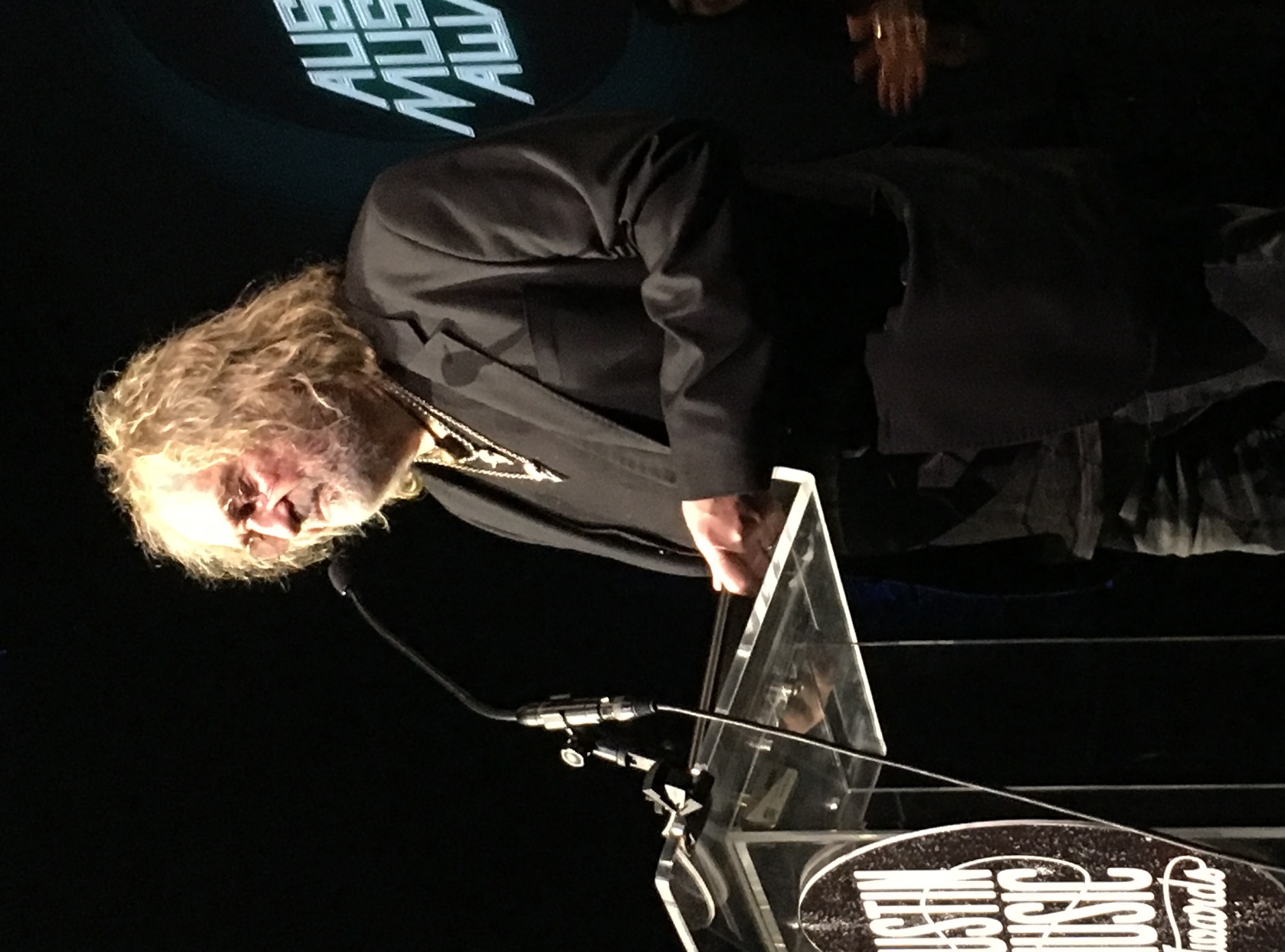 Ray Wylie Hubbard stands at a clear lectern in a black jacket, grey hair loose to his shoulders, speaking into a microphone at the Austin Music Awards.