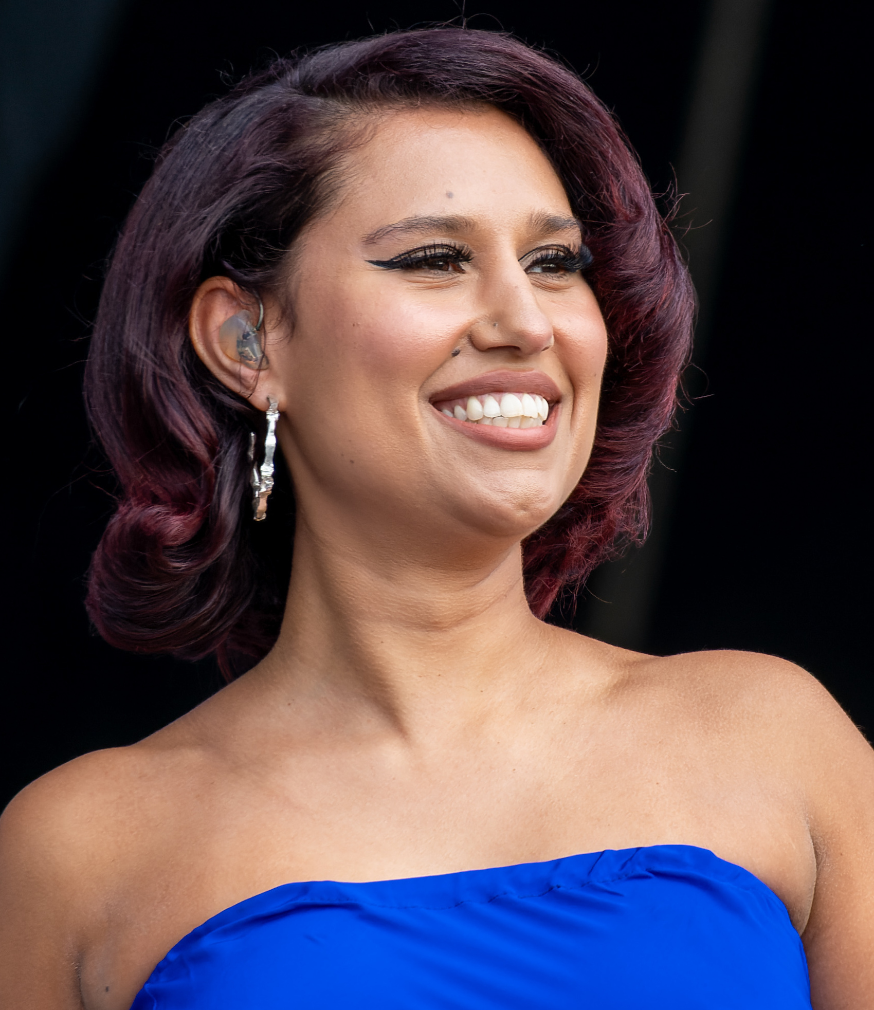 RAYE smiles off camera in a close portrait, her dark burgundy hair set in waves, wearing hoop earrings, an in-ear monitor and a blue strapless top.