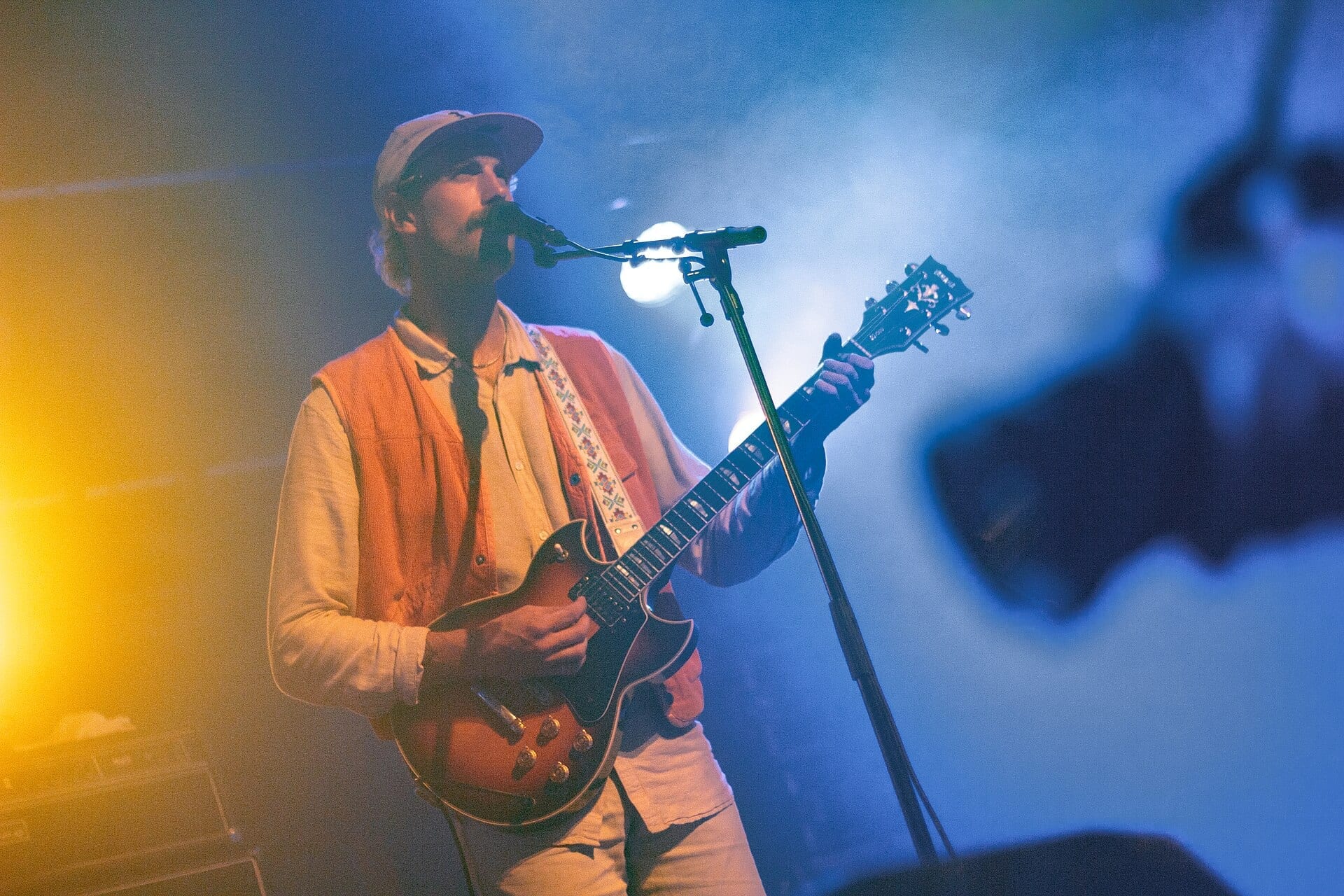 Rayland Baxter sings into a stand microphone in a pale cap and cream shirt, playing a sunburst electric guitar in warm and blue stage haze.