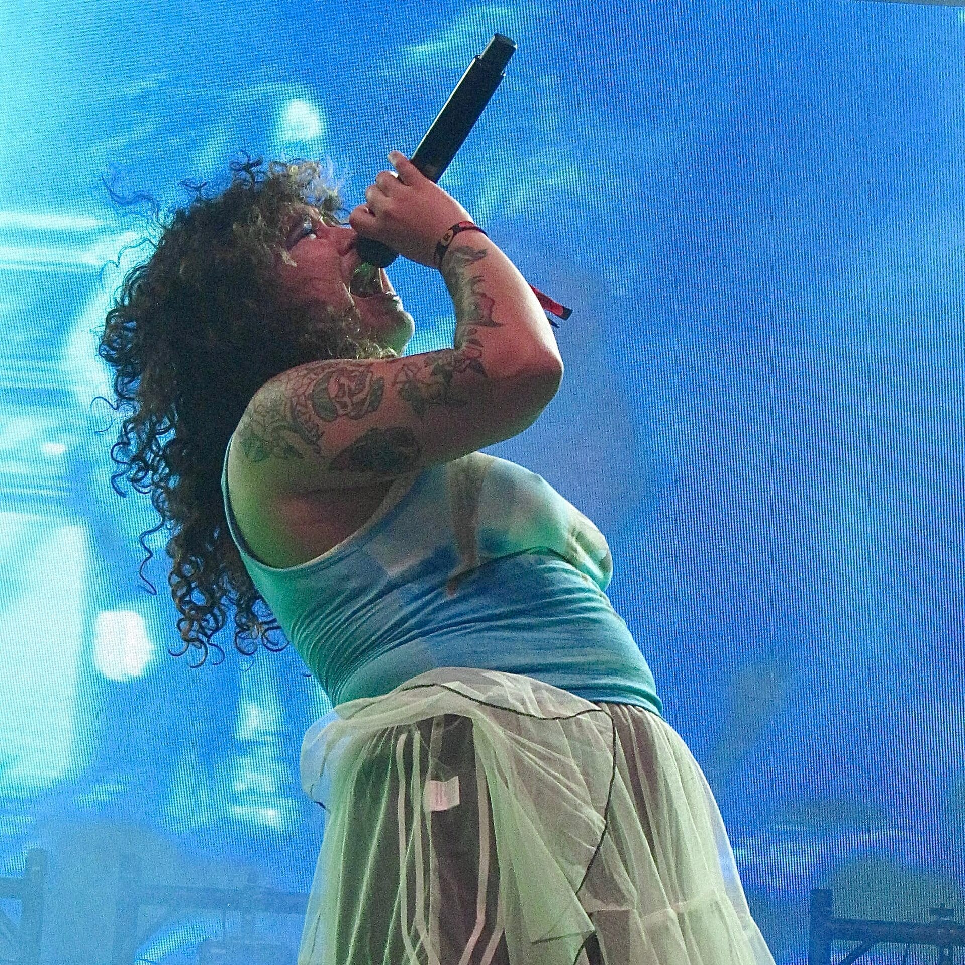 Remi Wolf, curly-haired and tattooed, sings with her head tilted back and mic raised, silhouetted against a blue screen.