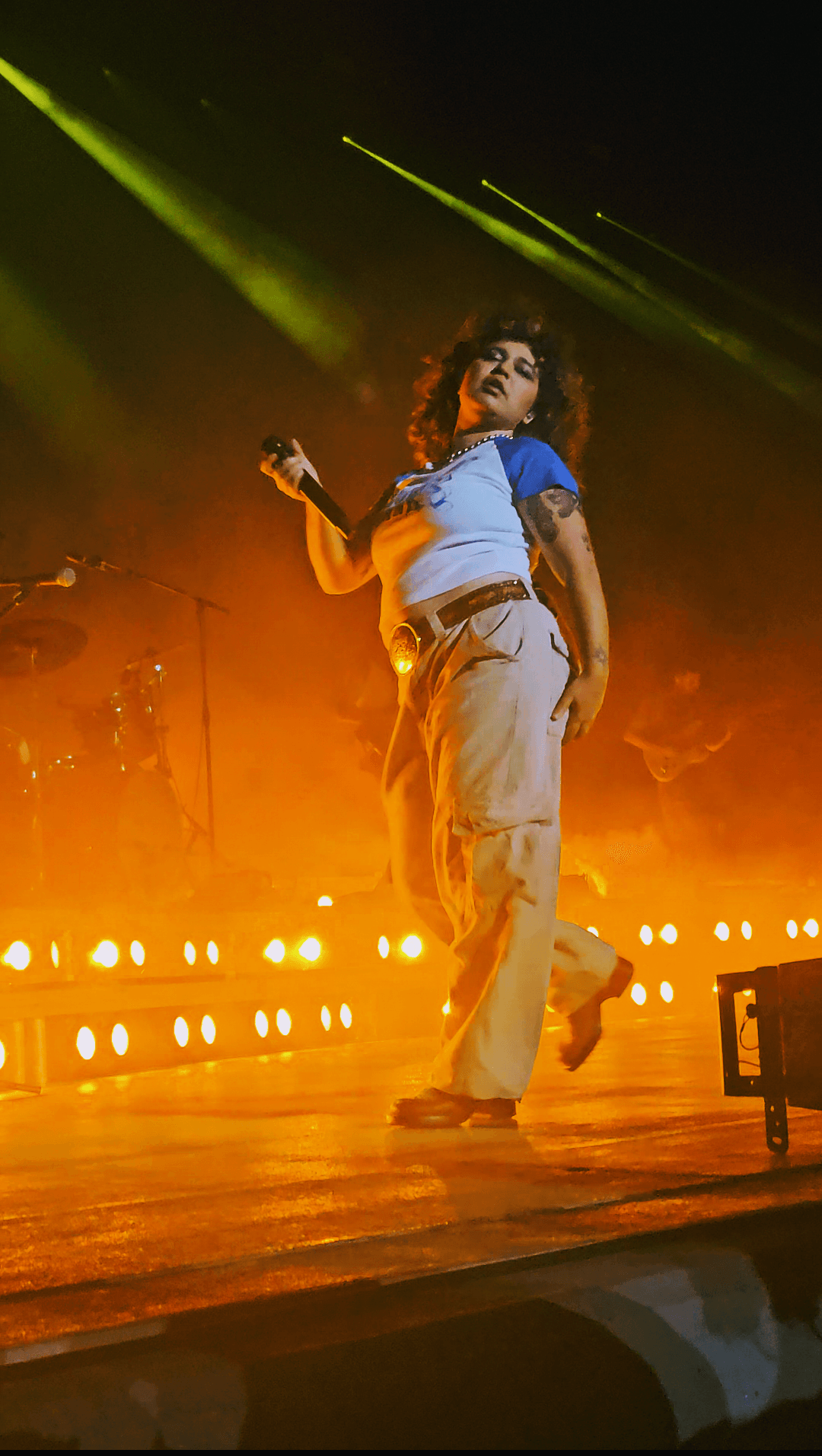 Remi Wolf, with curly dark hair and a baseball-style top, holds a microphone mid-stride on a stage bathed in orange light and green laser beams.