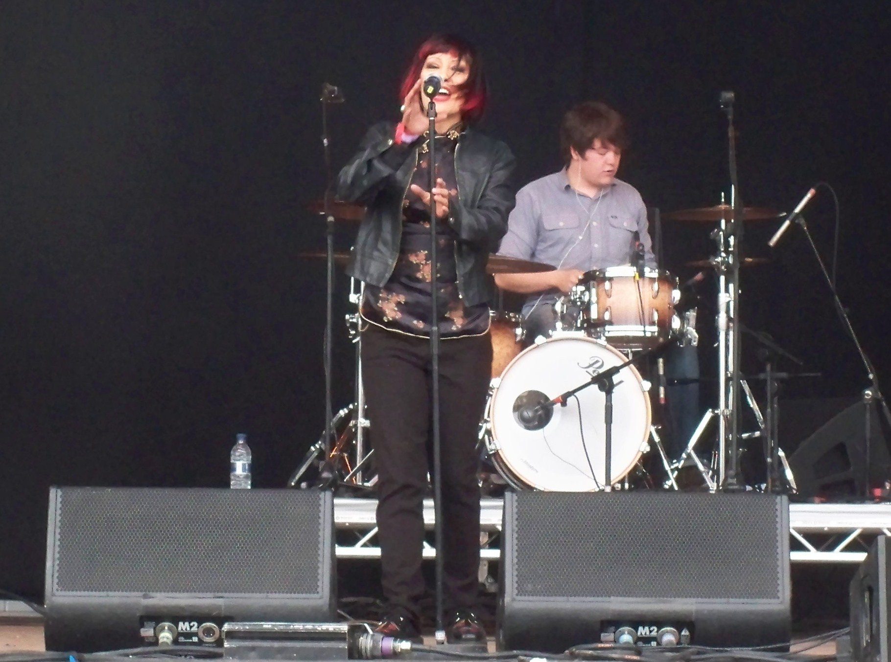 Republica's singer claps at a microphone stand in a black leather jacket, red streaks in her dark hair, with the drummer playing behind her on an outdoor festival stage.
