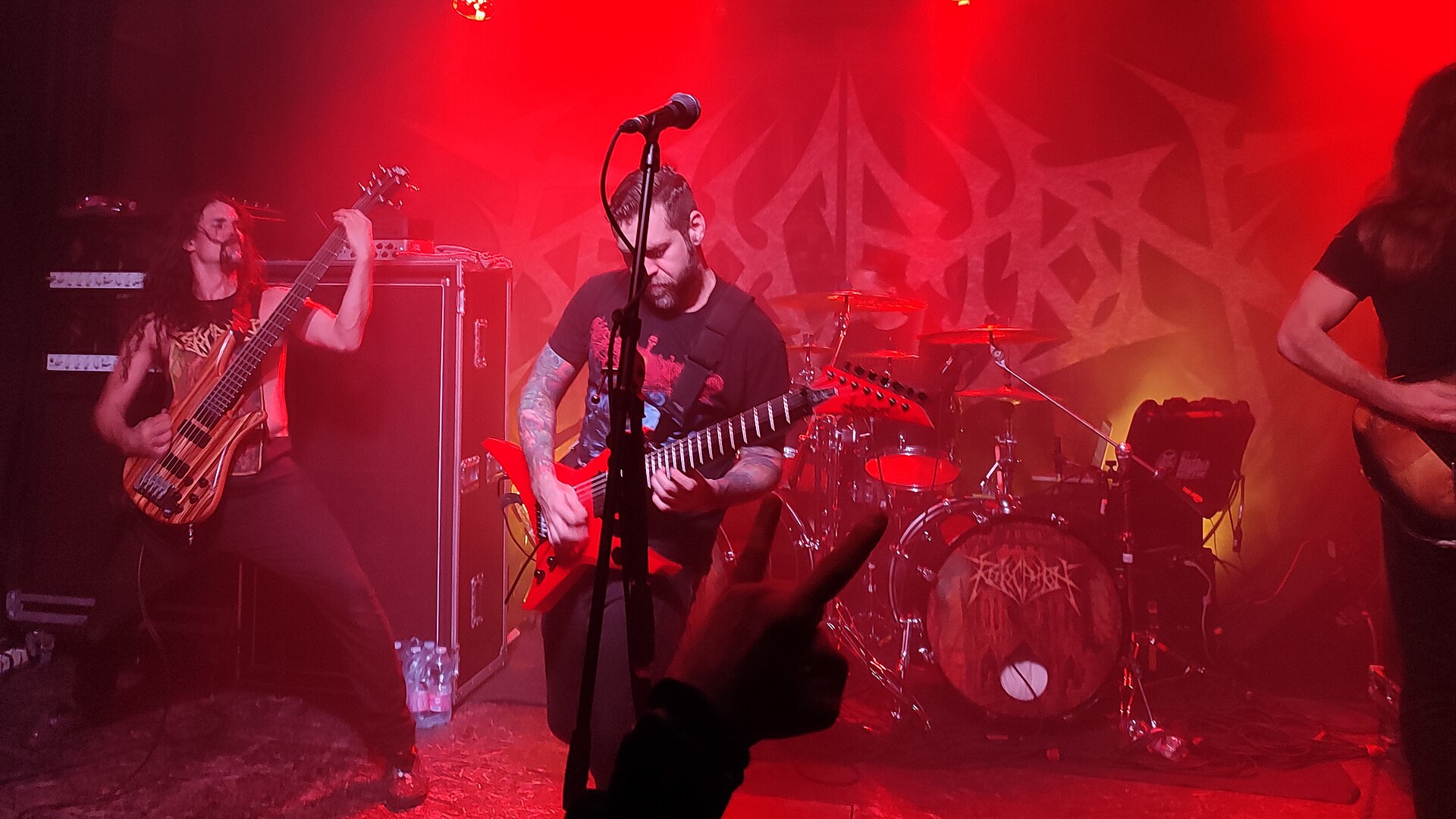 Revocation play a club stage flooded in red light, the bearded singer centre on a red flying-V, a long-haired bassist to his left and the drum kit behind carrying the band's logo.