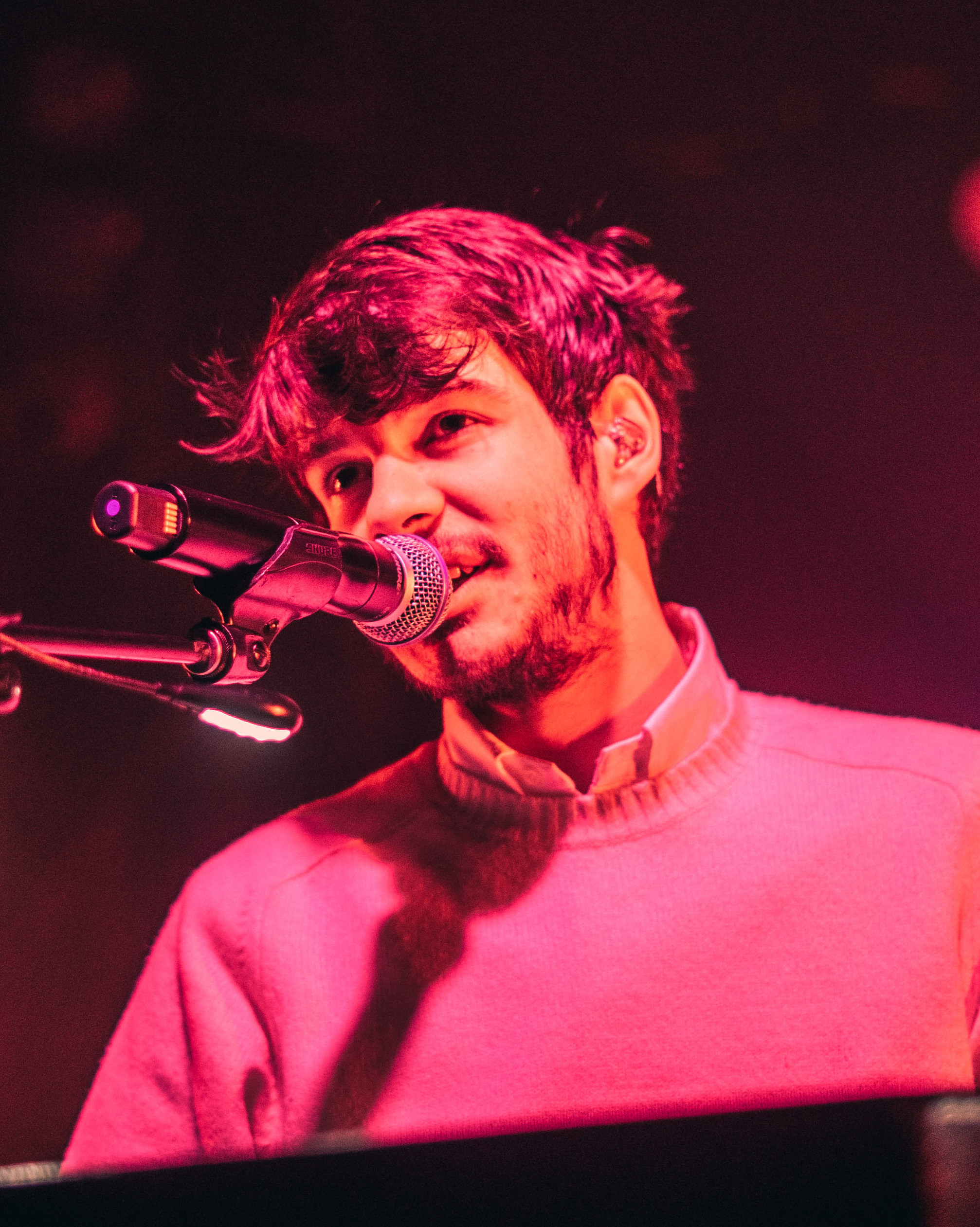 Rex Orange County sings into a microphone behind a keyboard in a pink crewneck jumper, washed in red stage light.