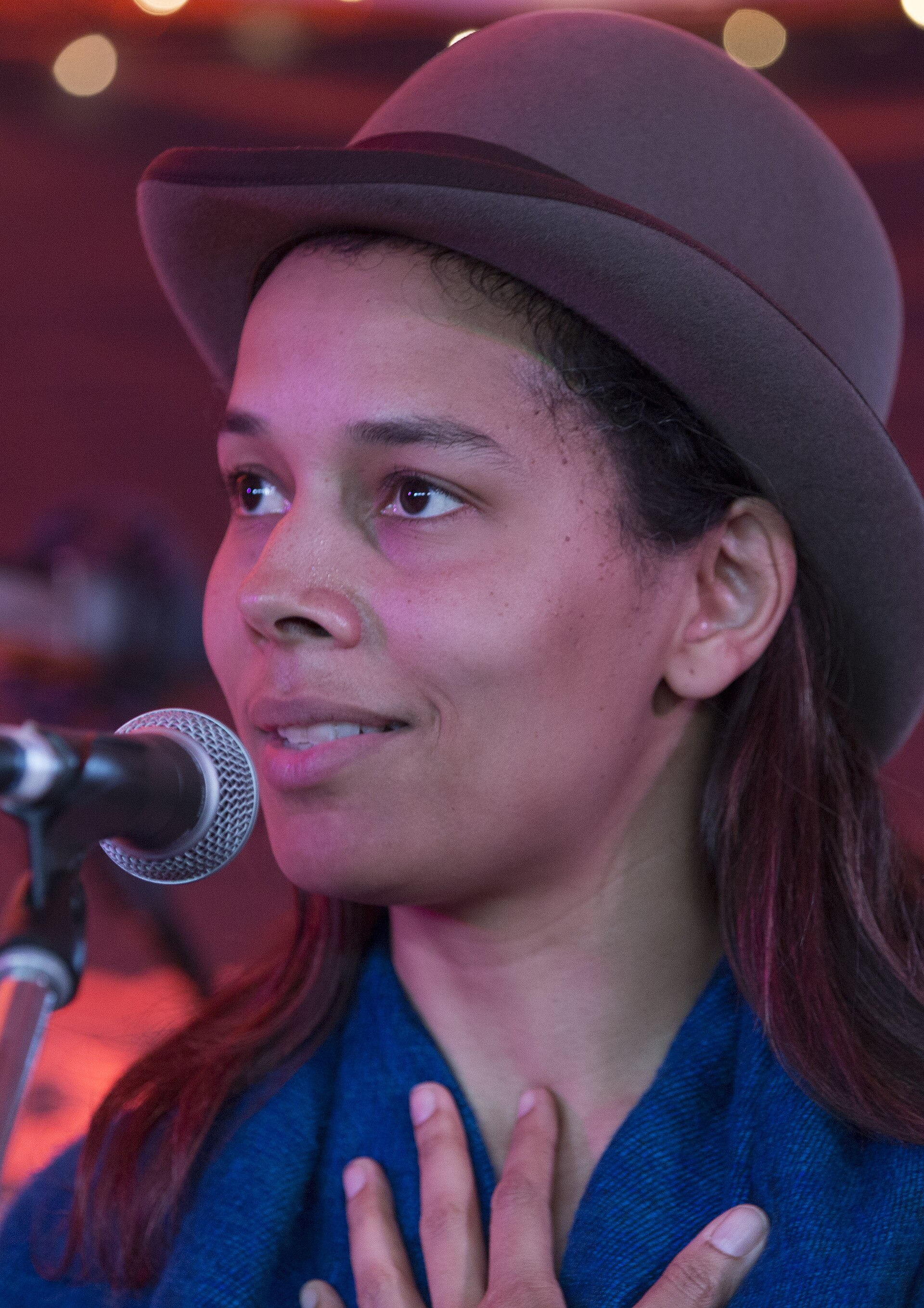 A woman in a felt bowler hat and a blue wrap stands at a microphone with one hand to her chest, looking off to the side under warm stage light
