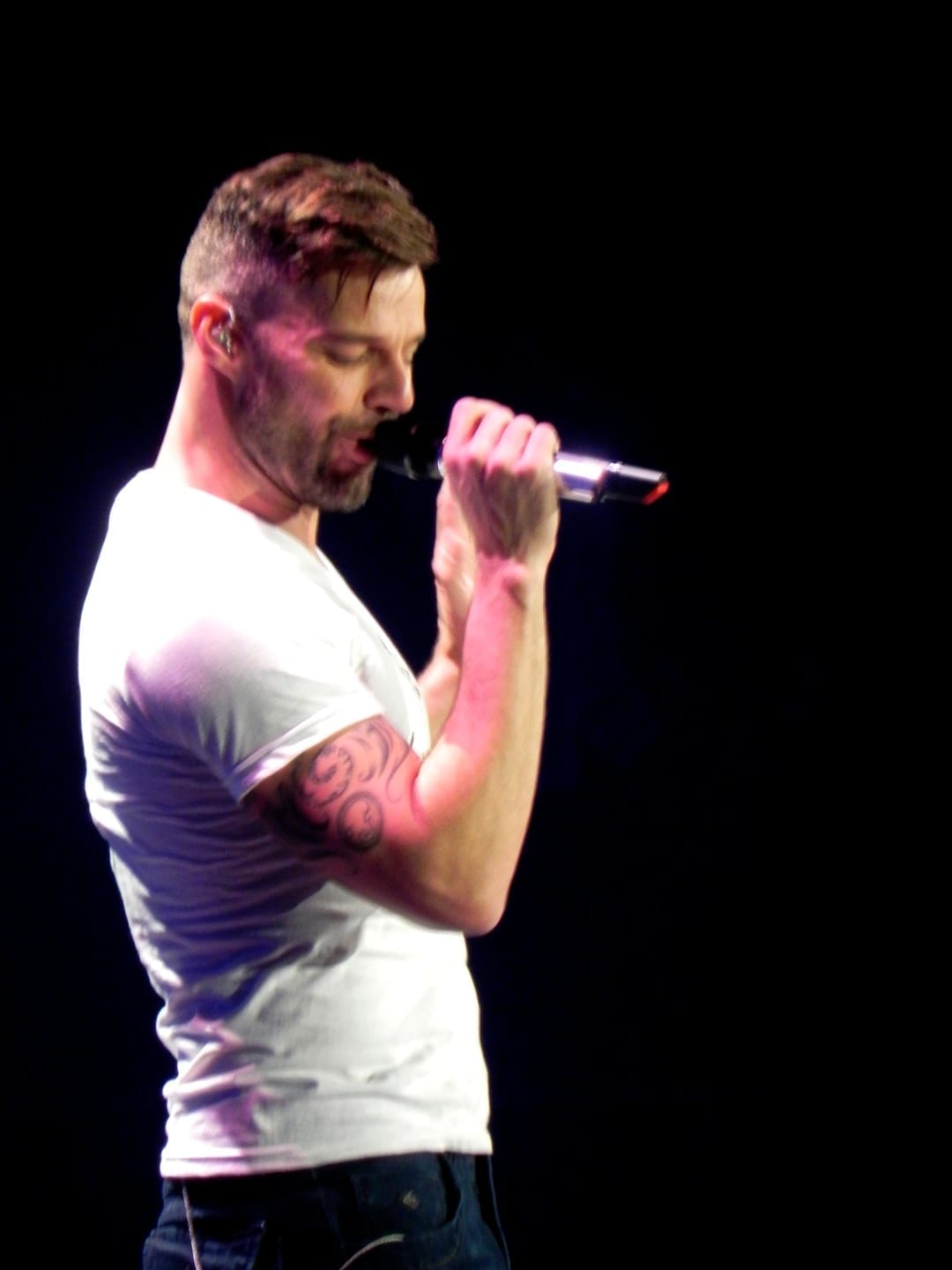 Ricky Martin, in a white t-shirt with a tattooed forearm, sings into a handheld microphone against a dark background.