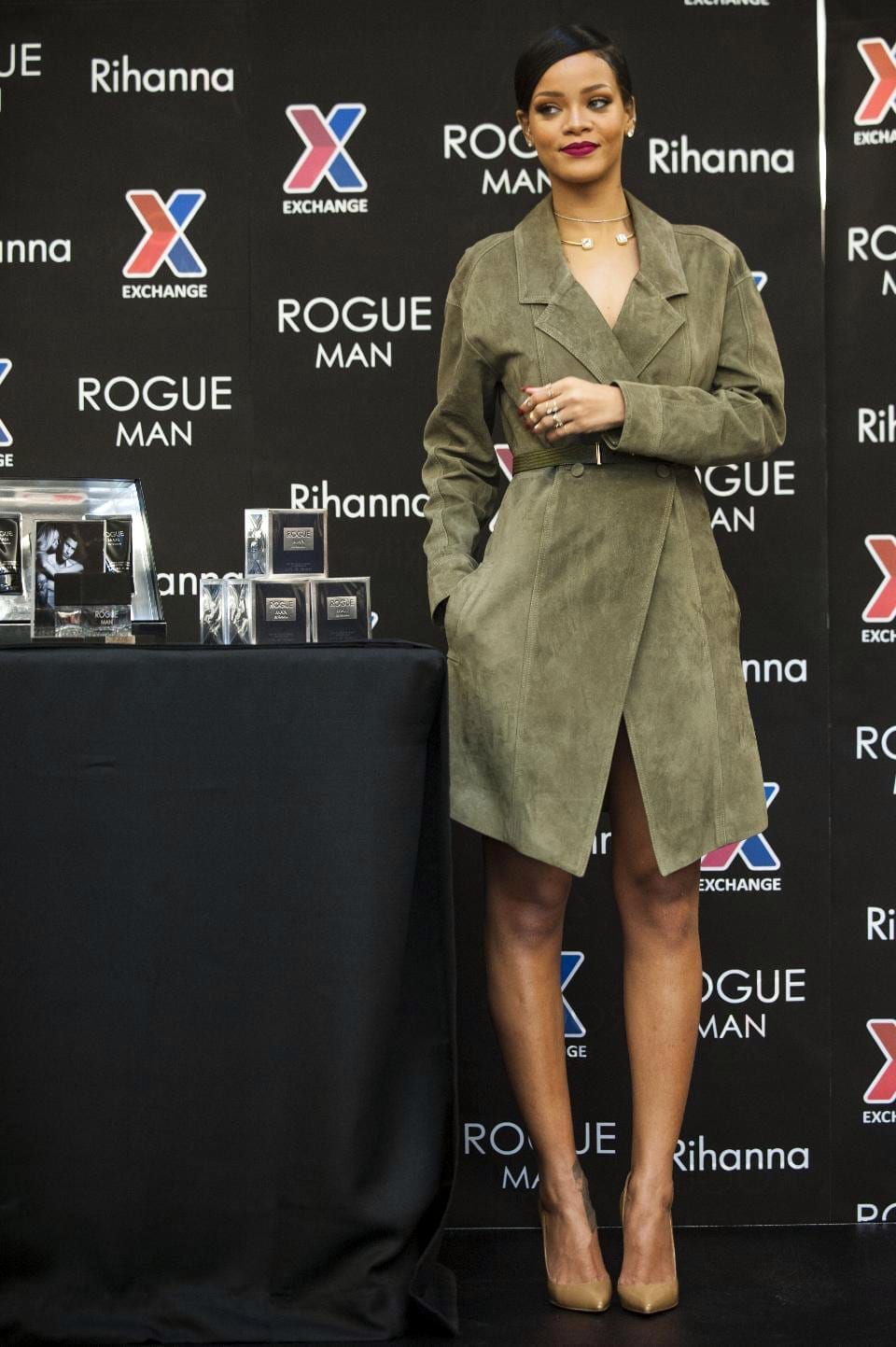 Rihanna stands in an olive suede trench coat in front of a step-and-repeat backdrop bearing her name.