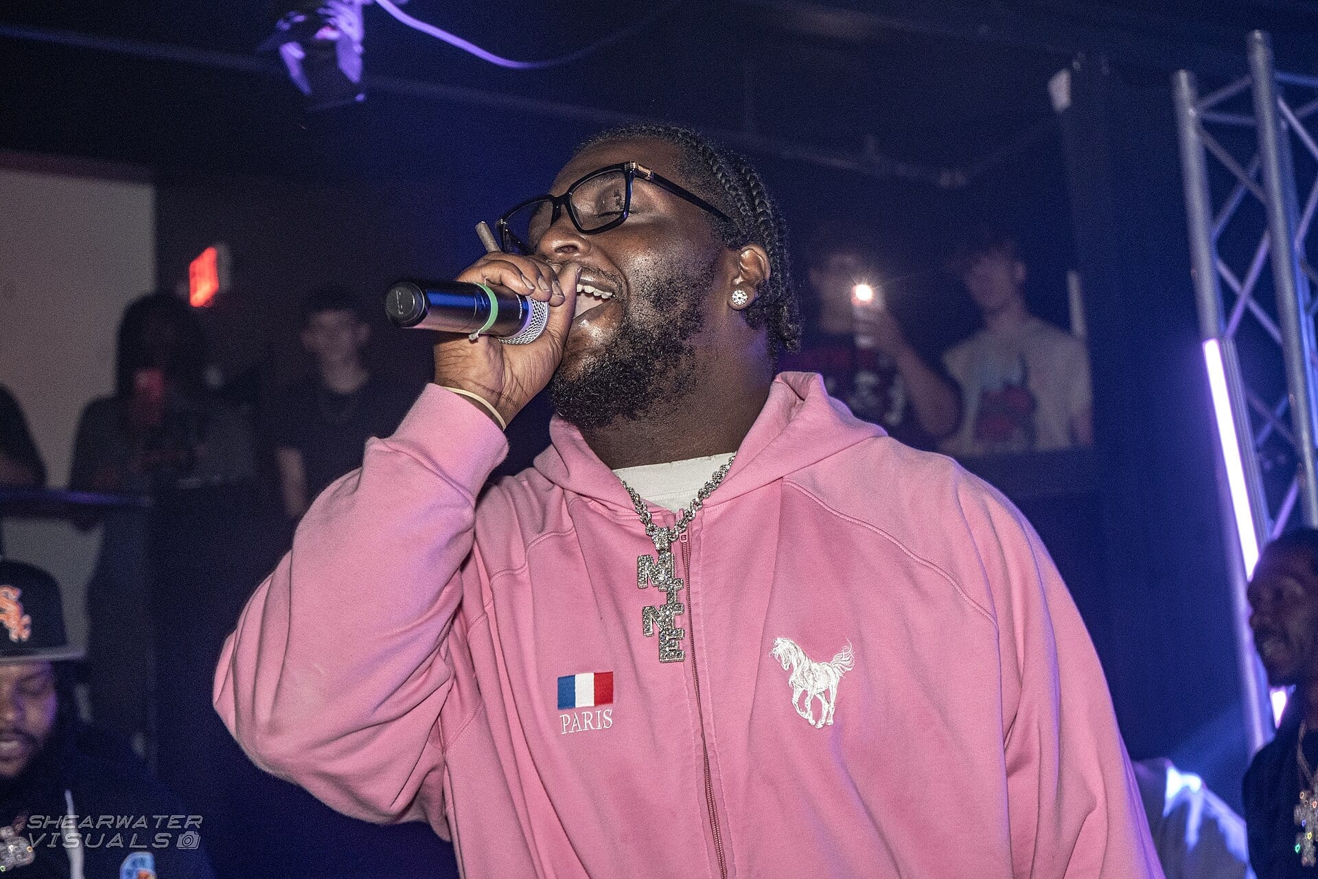 Rio Da Yung OG raps into a handheld microphone in a pink zip hoodie and glasses, a diamond pendant at his chest and phones held up in the crowd behind him.