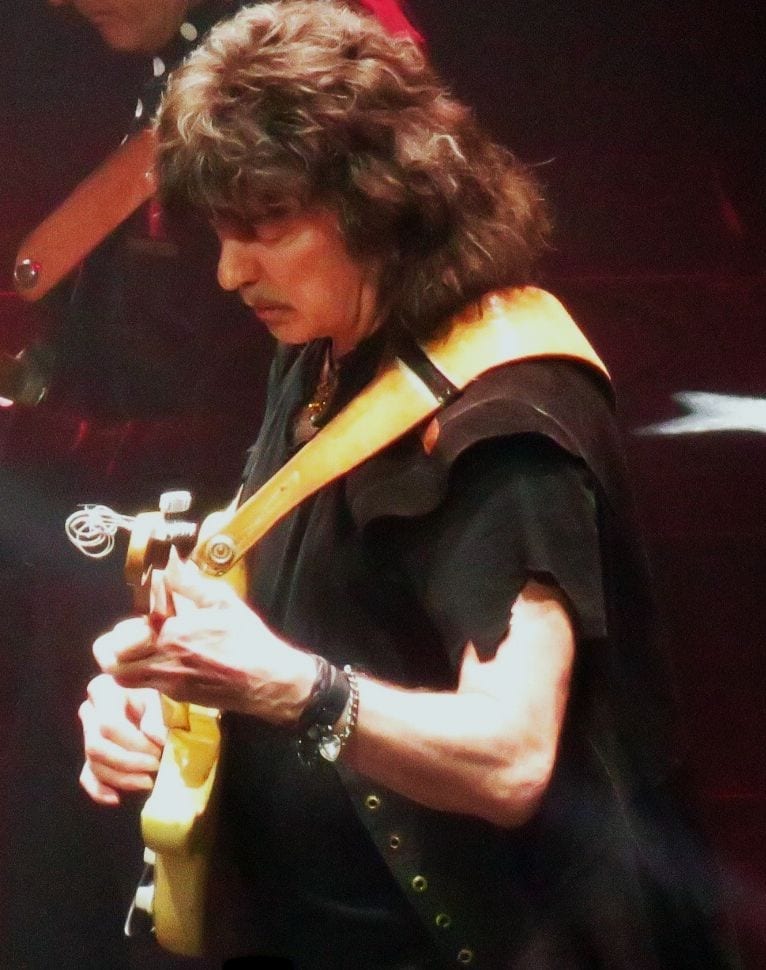 Ritchie Blackmore, in profile with tousled shoulder-length hair, plays a yellow electric guitar on a dark, red-lit stage.