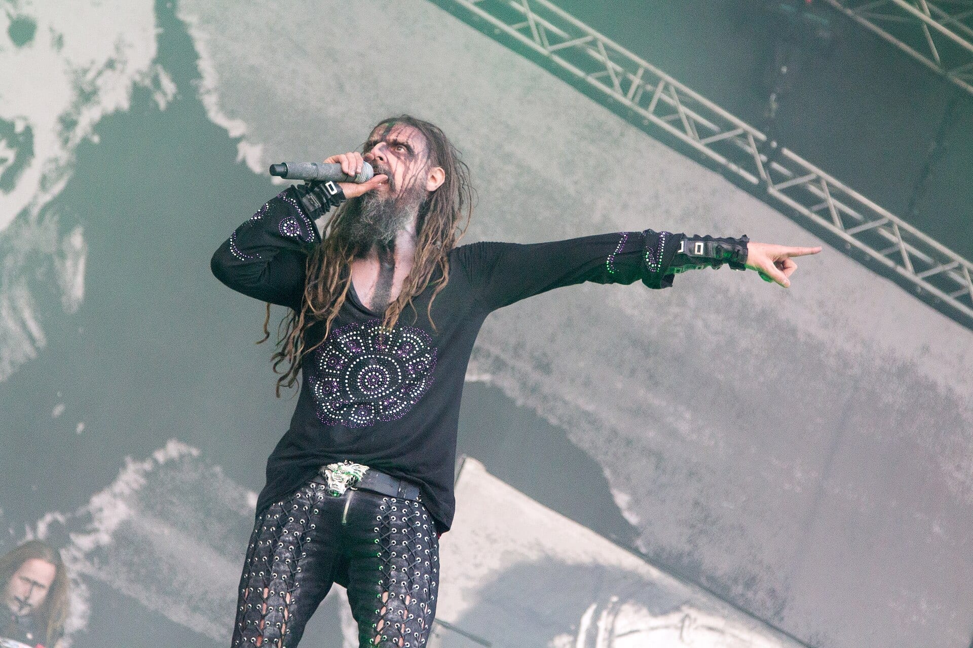 Rob Zombie sings into a microphone and points off stage, wearing black stage clothes with his long dreadlocks loose.