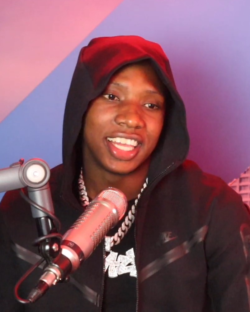 Rob49 smiles mid-sentence in a red and black hoodie and a diamond chain, behind a studio microphone against a pink and purple wall.