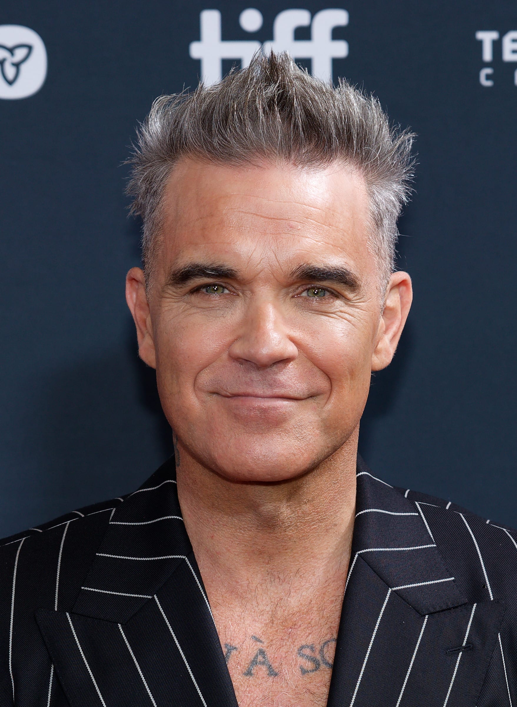 Robbie Williams smiles at the camera in close-up on a red carpet, wearing a black pinstripe suit jacket, with TIFF festival signage visible behind him.