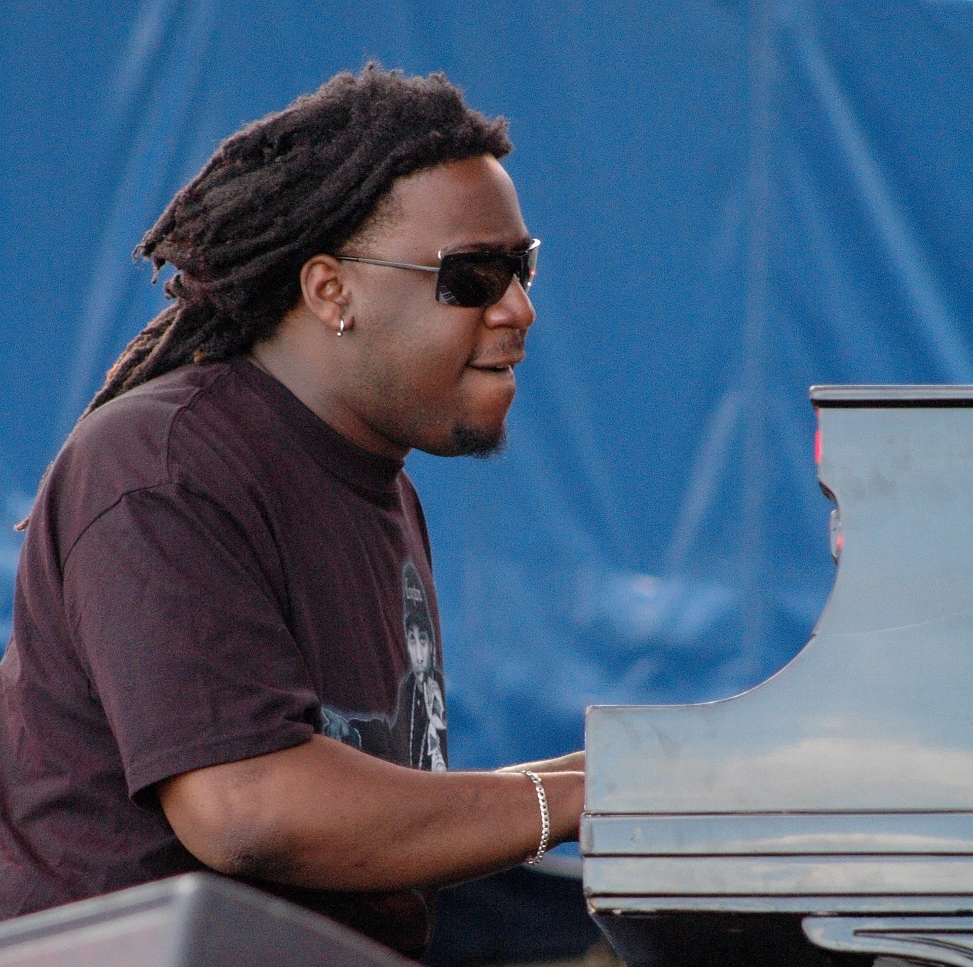 Robert Glasper, wearing sunglasses with dreadlocks pulled back, plays a grand piano in profile against a blue backdrop.