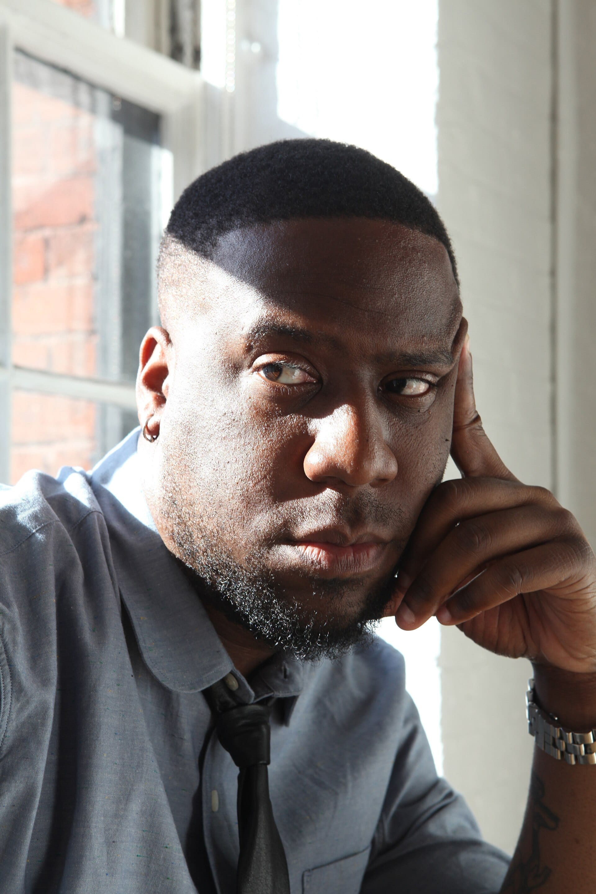 Robert Glasper rests his chin on his hand by a bright window, wearing a grey shirt and loosened black tie, looking off to the side.