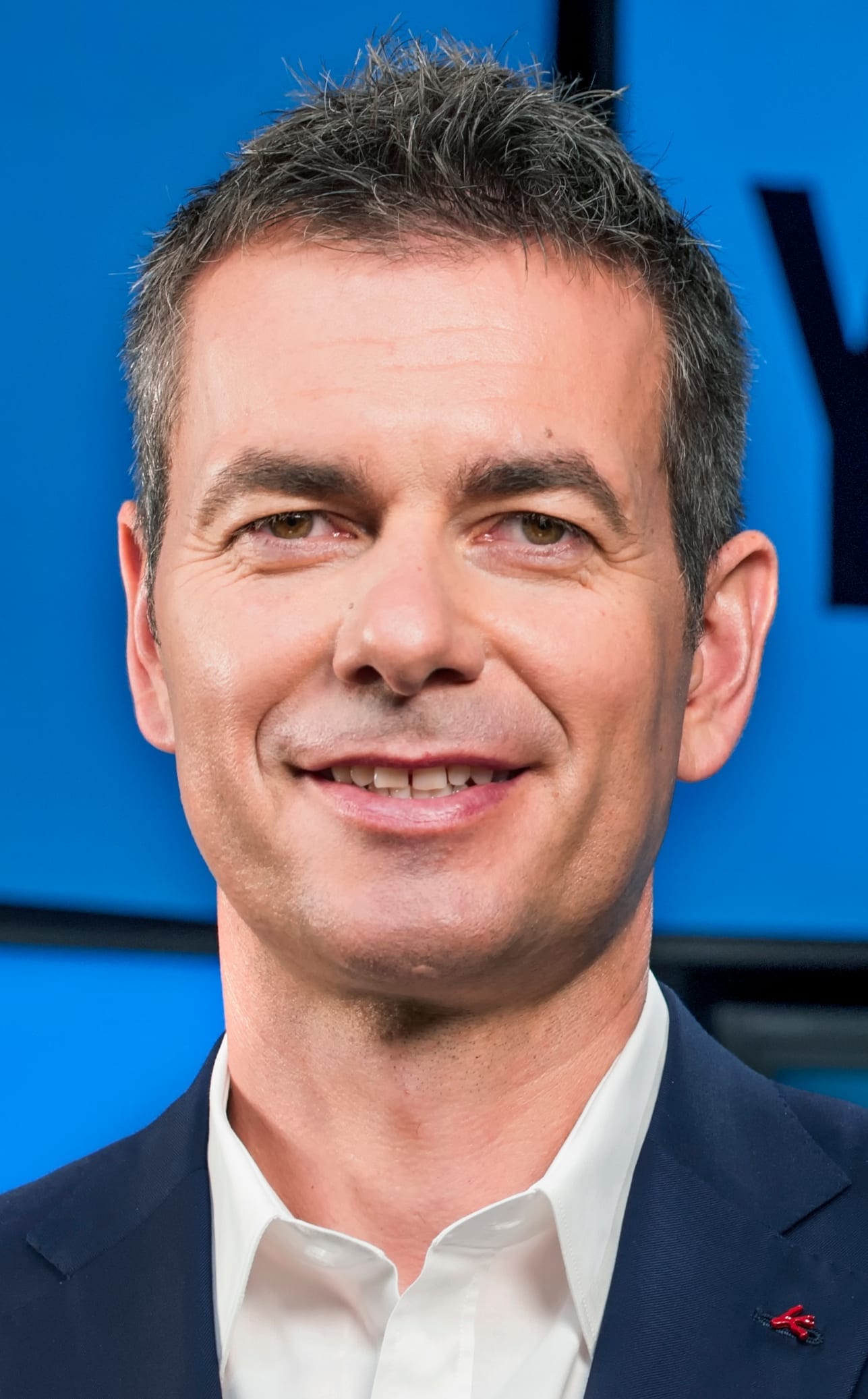 Robert Kyncl, in a dark suit and white open-collar shirt, smiles in a close-up headshot against a blue backdrop.