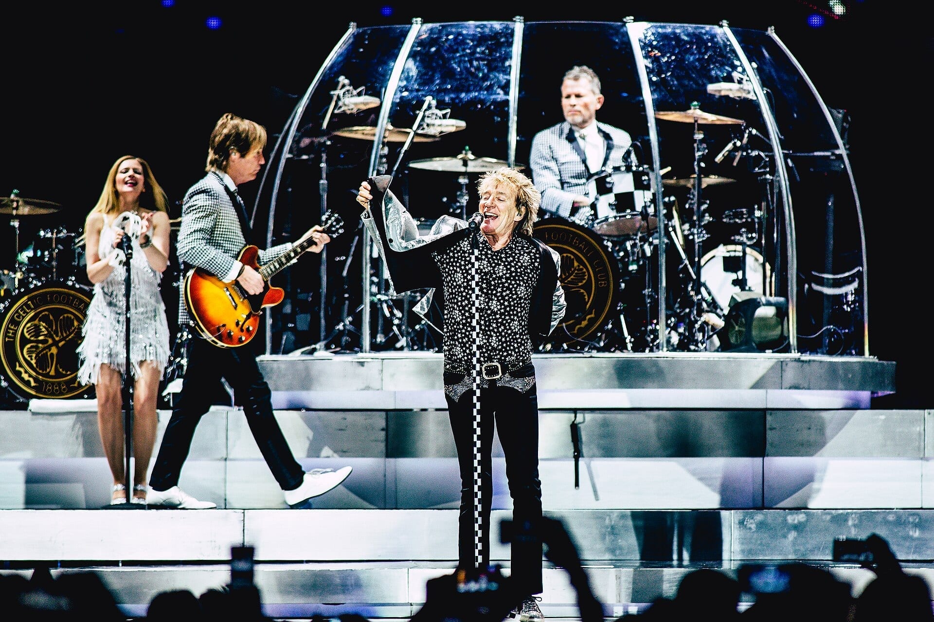 Rod Stewart sings into a microphone on a bright arena stage, wearing a star-patterned top and checkered pants, with a guitarist, a backing vocalist, and a drummer on a round glass-paneled riser behind him.
