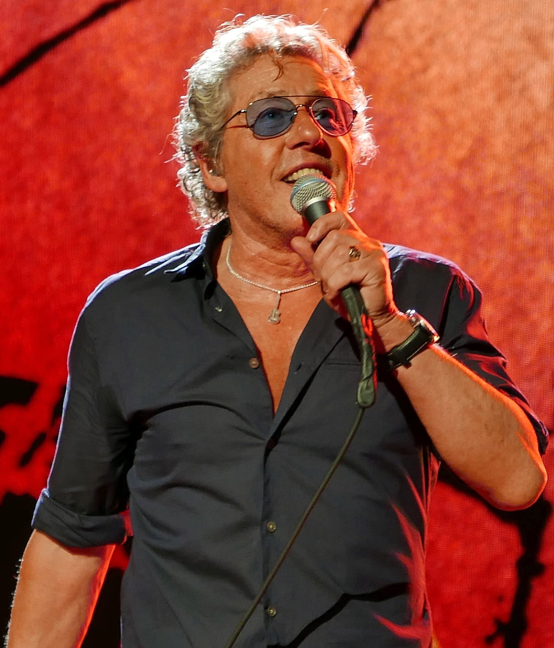 Roger Daltrey sings into a handheld microphone in tinted round sunglasses and an open dark shirt, his white curls lit orange against a red backdrop.