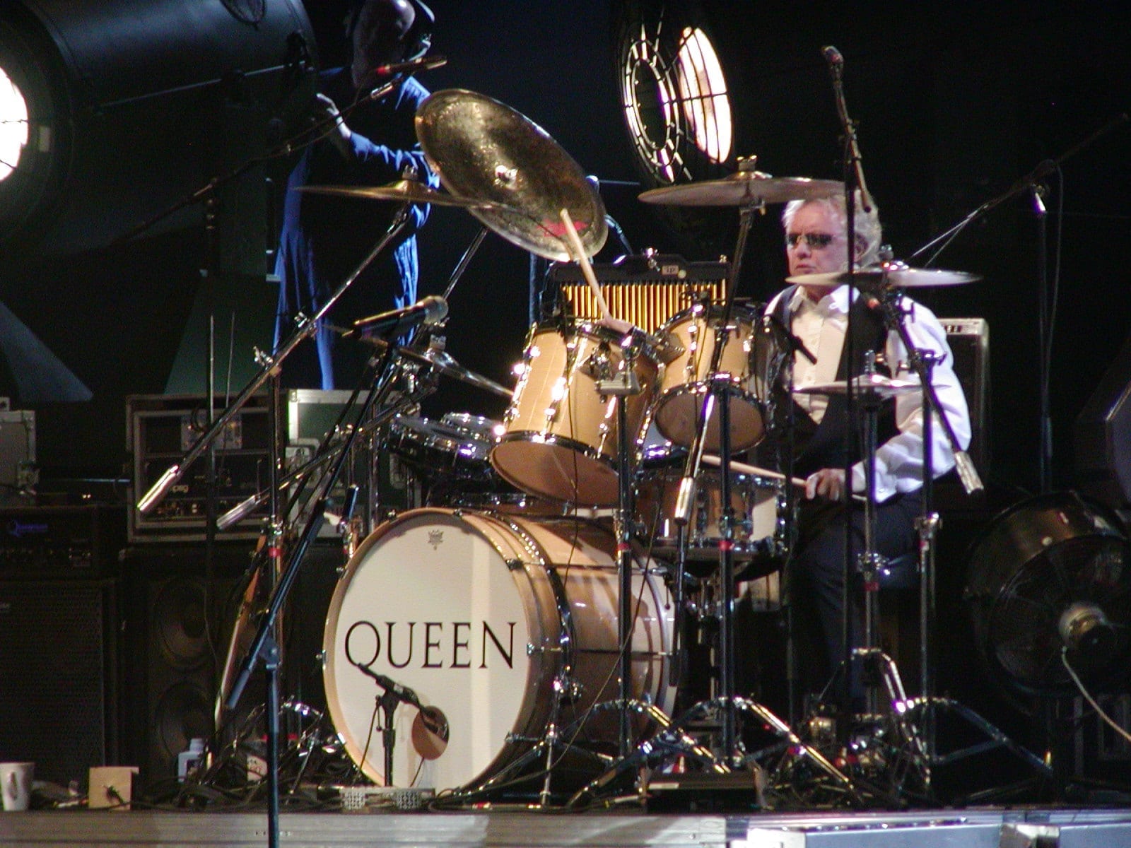 Roger Taylor, wearing sunglasses, plays a drum kit with a bass drum head reading "Queen" on a dark stage.