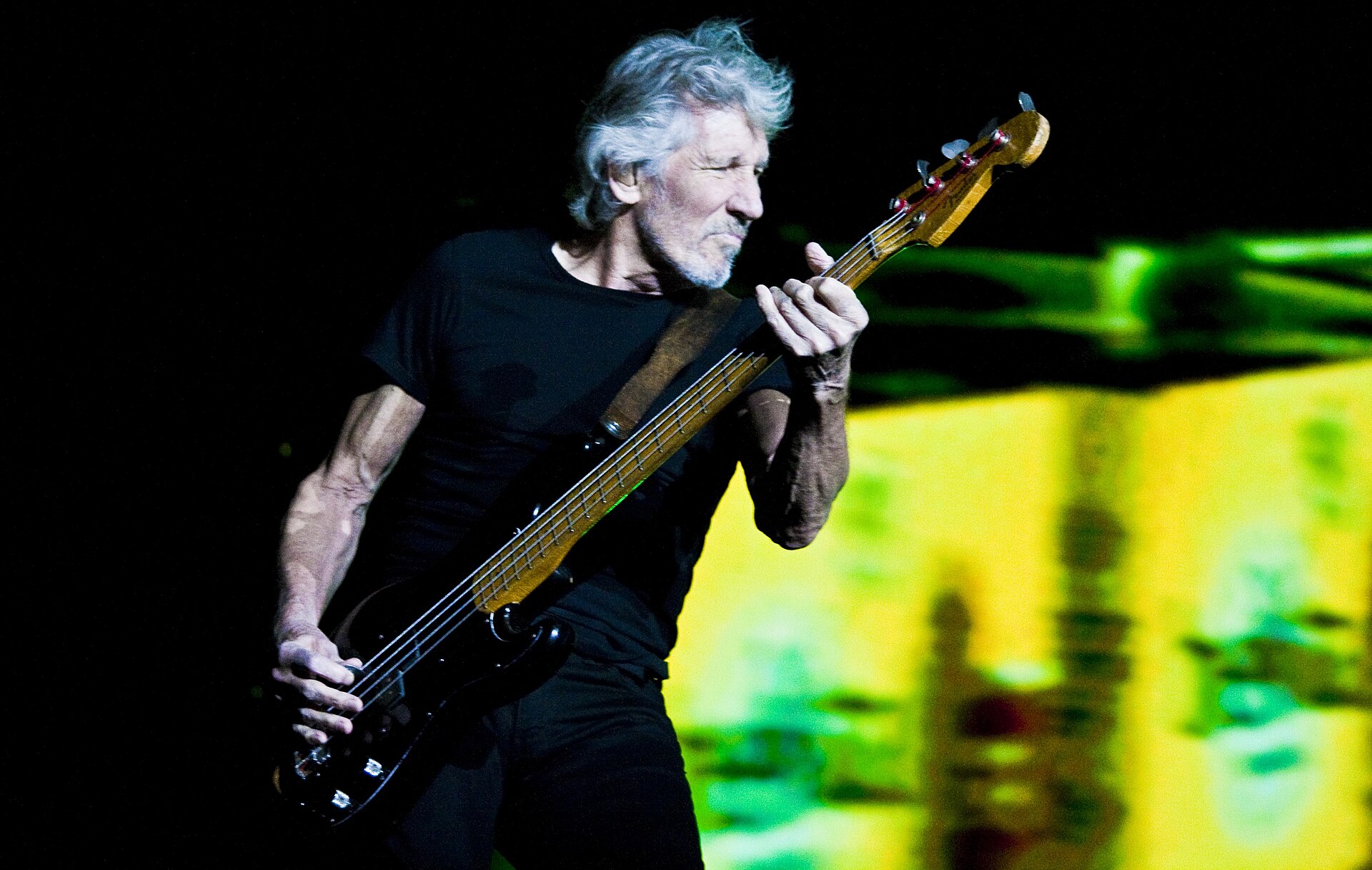 Roger Waters plays a worn bass guitar in a plain black T-shirt, white hair pushed back, a green and yellow projection behind him.