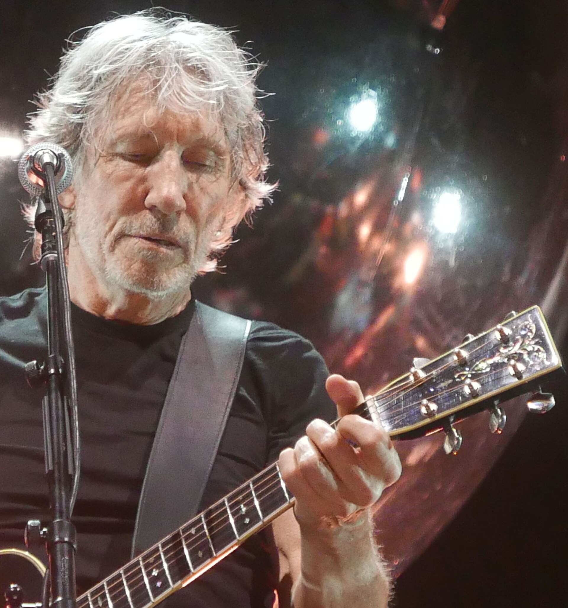 Close-up of Roger Waters on stage, white hair backlit by stage lamps, eyes down on the fretboard of an acoustic guitar with a microphone at his mouth.