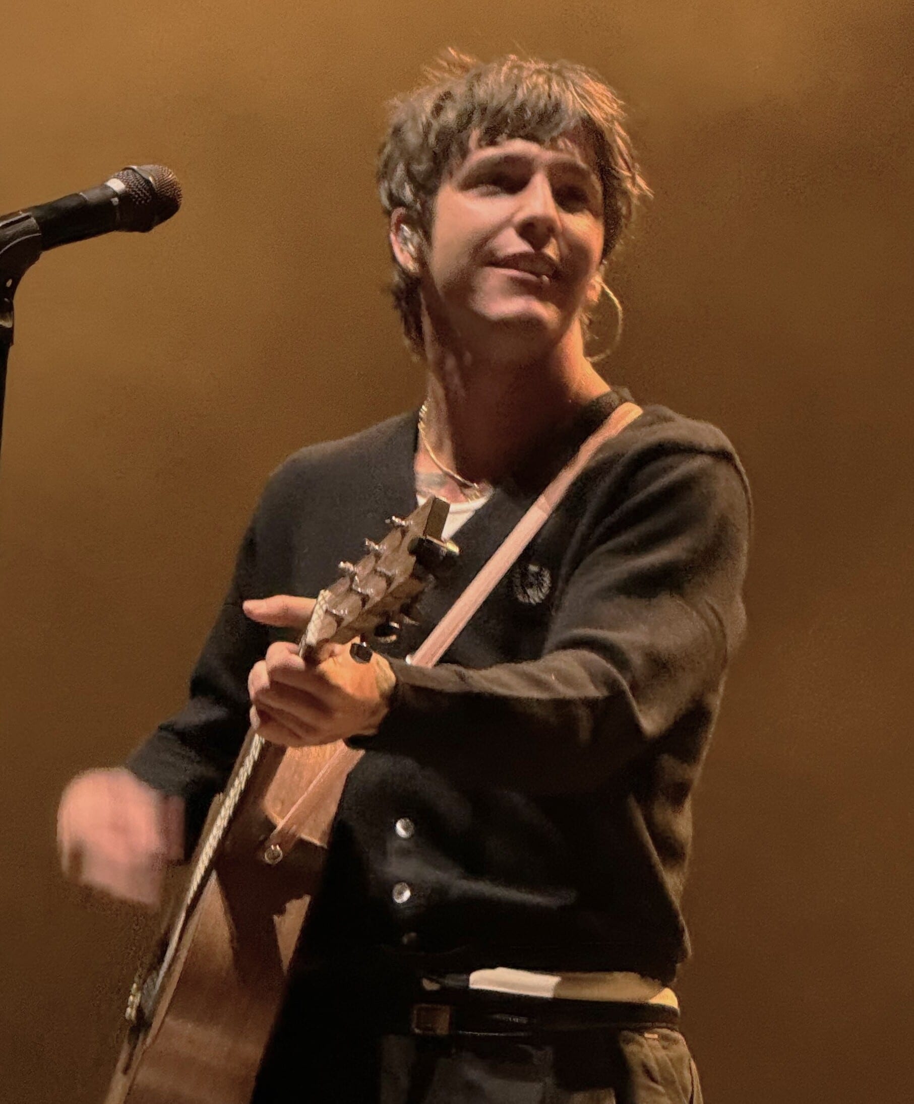 Role Model (Tucker Pillsbury) smiles while holding a guitar neck on stage, wearing a black cardigan and hoop earring near a microphone stand.