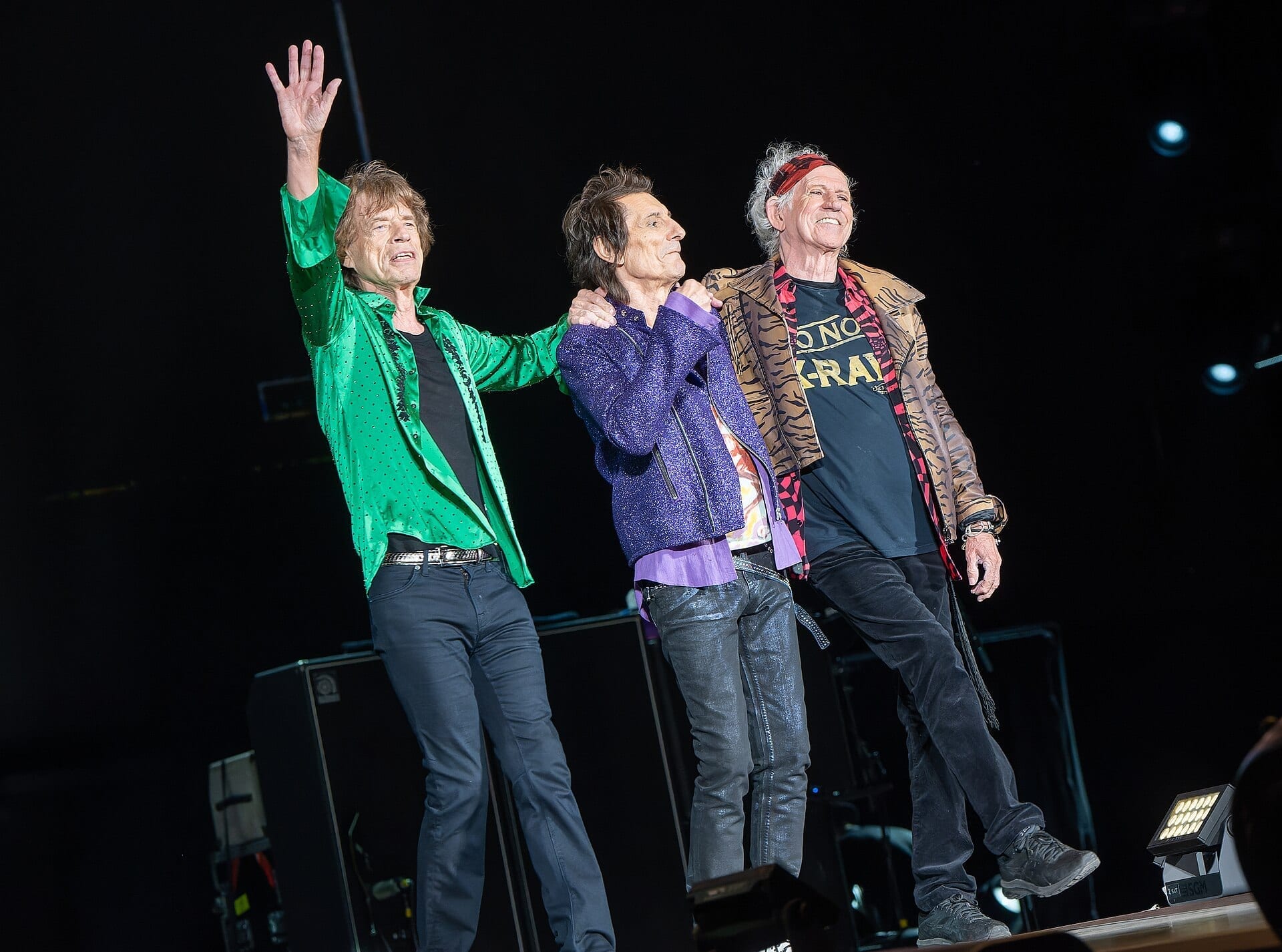 Mick Jagger, Ronnie Wood and Keith Richards stand arm in arm on stage taking a bow, Jagger waving to the crowd.