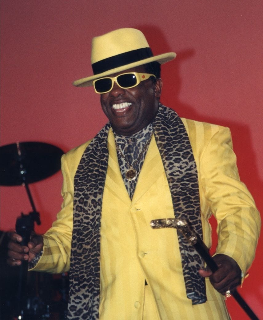 Ronald Isley smiles while wearing a yellow suit, a matching hat and sunglasses, a leopard-print scarf, and holding a gold-topped cane.