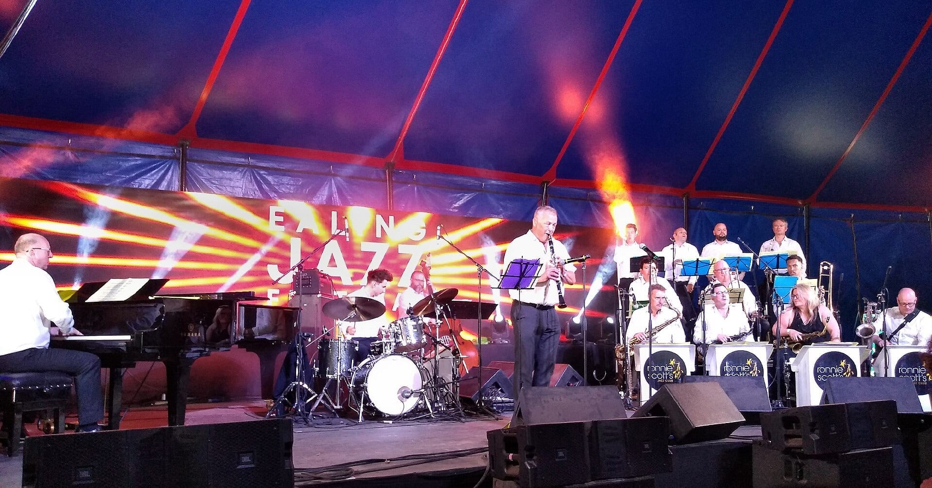 Ronnie Scott's Jazz Orchestra performs on a red and blue lit stage under a festival tent, with a pianist and drummer at left, a clarinet soloist at center, and a full horn section in white shirts behind them.