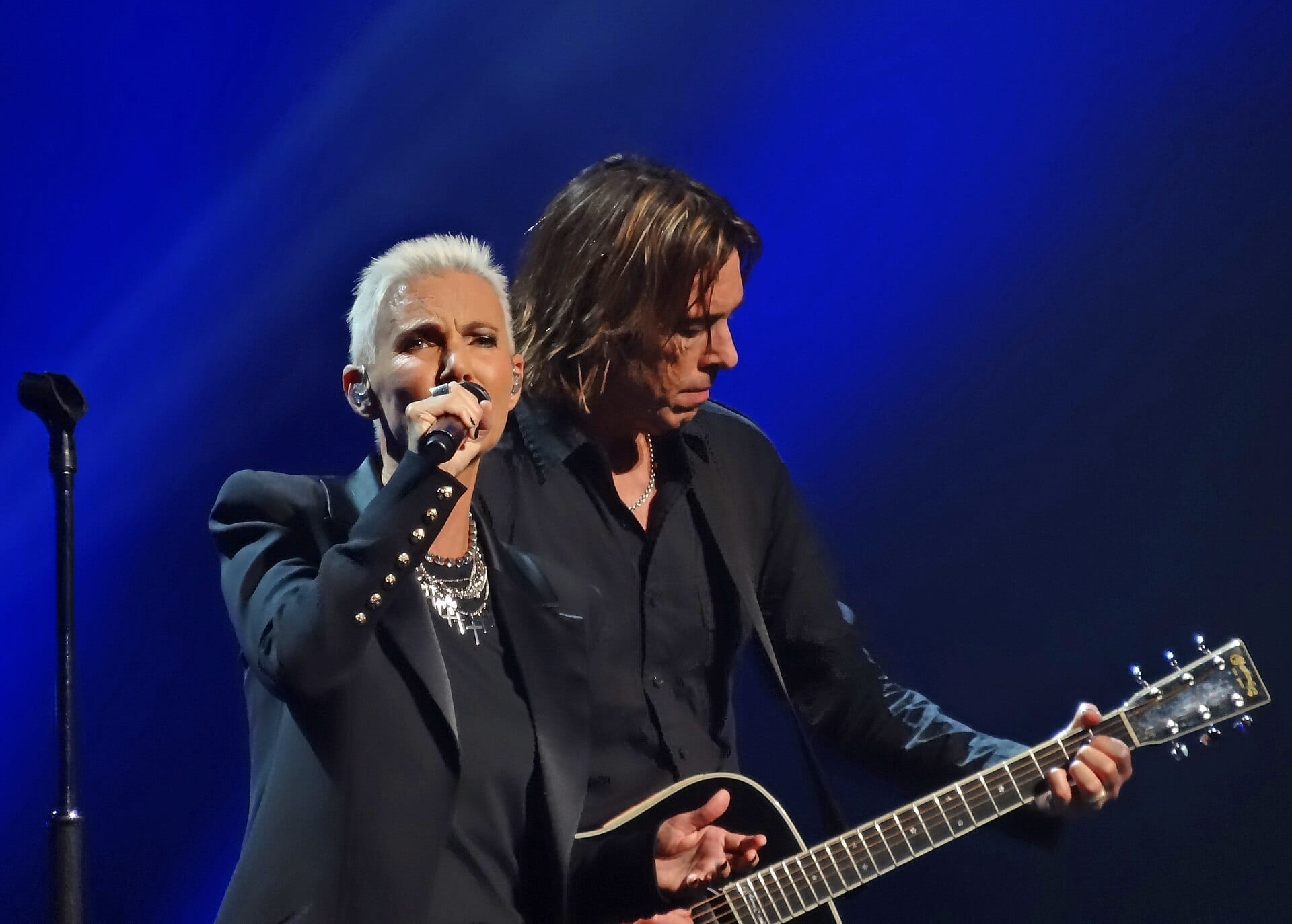 Marie Fredriksson sings into a microphone with short white hair and a studded black blazer, while Per Gessle plays an acoustic guitar beside her against a deep blue stage backdrop.