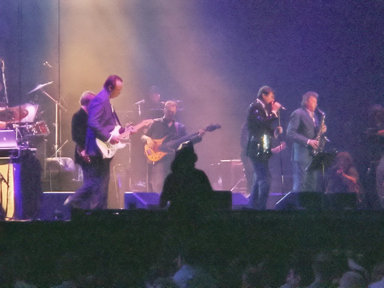 Roxy Music performing on stage, band members playing guitars and singing into microphones under purple and gold stage lighting, with the audience visible in the foreground.