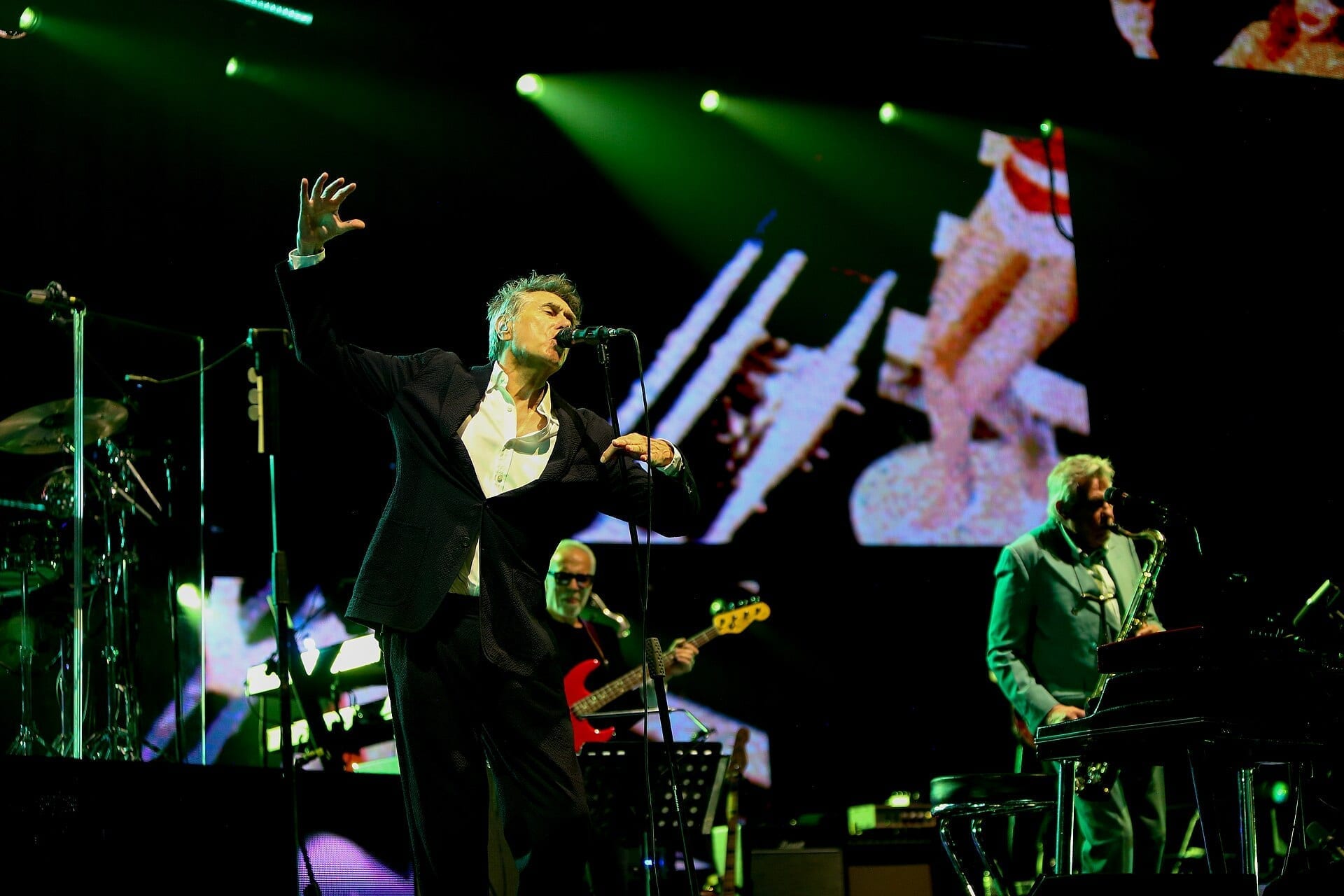 Bryan Ferry sings with one arm raised at a microphone stand, with a bassist and saxophonist performing behind him under green stage lights.
