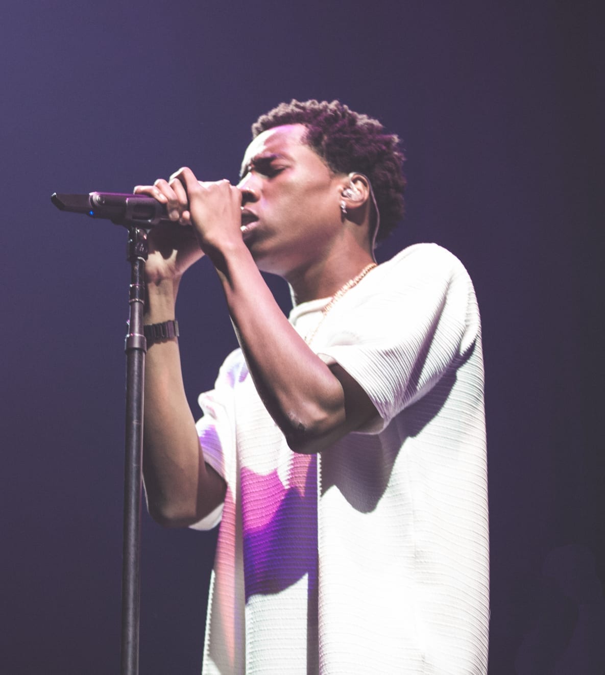 Roy Woods sings into a microphone in a loose white top and gold chain necklace, with his eyes closed under stage lighting.
