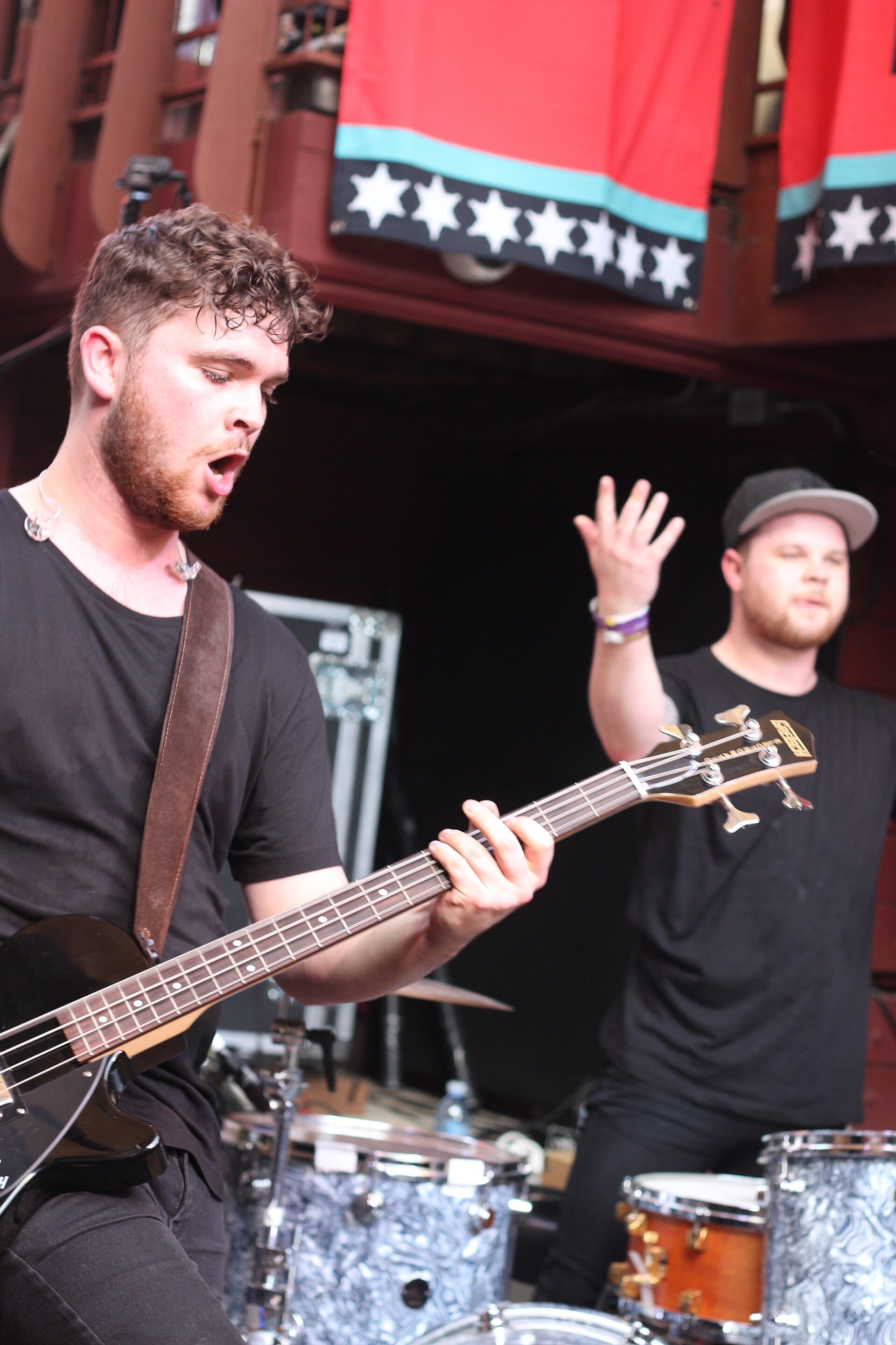 Royal Blood perform outdoors on stage: the bassist sings into the air with his mouth open while playing a black bass guitar, and the drummer stands behind the kit raising one hand.