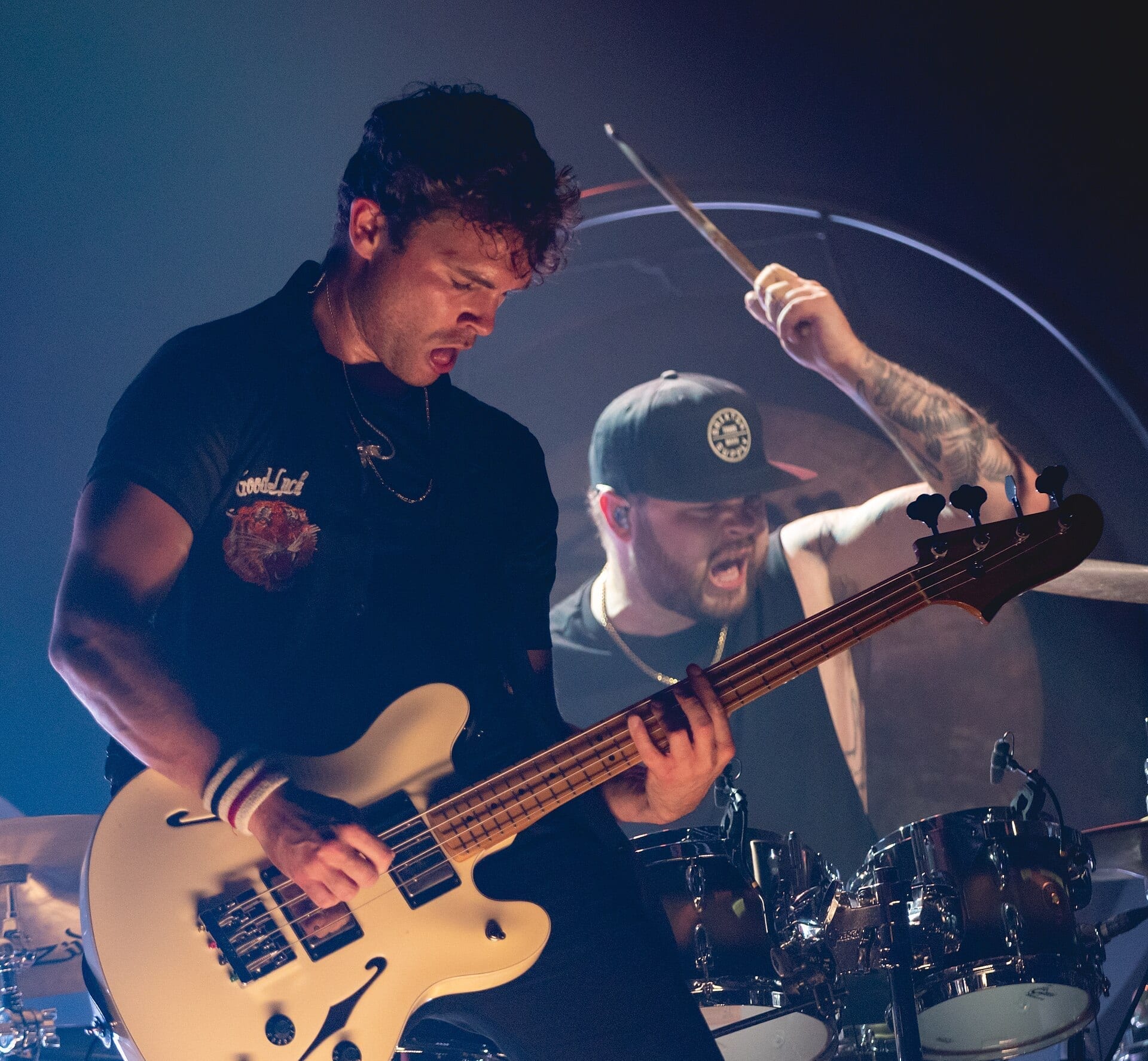 Royal Blood mid-song: Mike Kerr shouts while playing a cream hollow-body bass in the foreground while drummer Ben Thatcher, in a dark cap, swings a stick behind him.