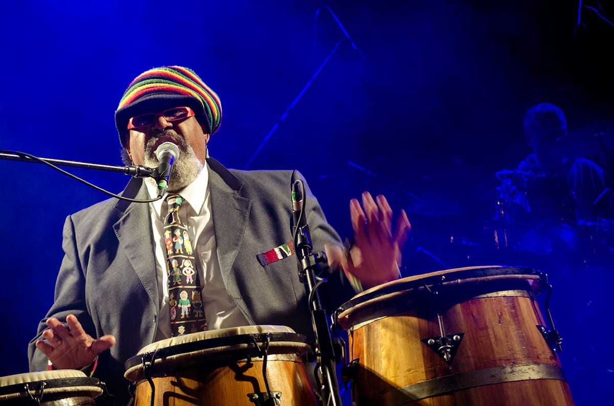 Ruben Rada sings at a microphone behind a set of tumbadora drums, hands raised mid-beat, wearing a striped knit cap, red glasses, a grey jacket and a patterned tie.