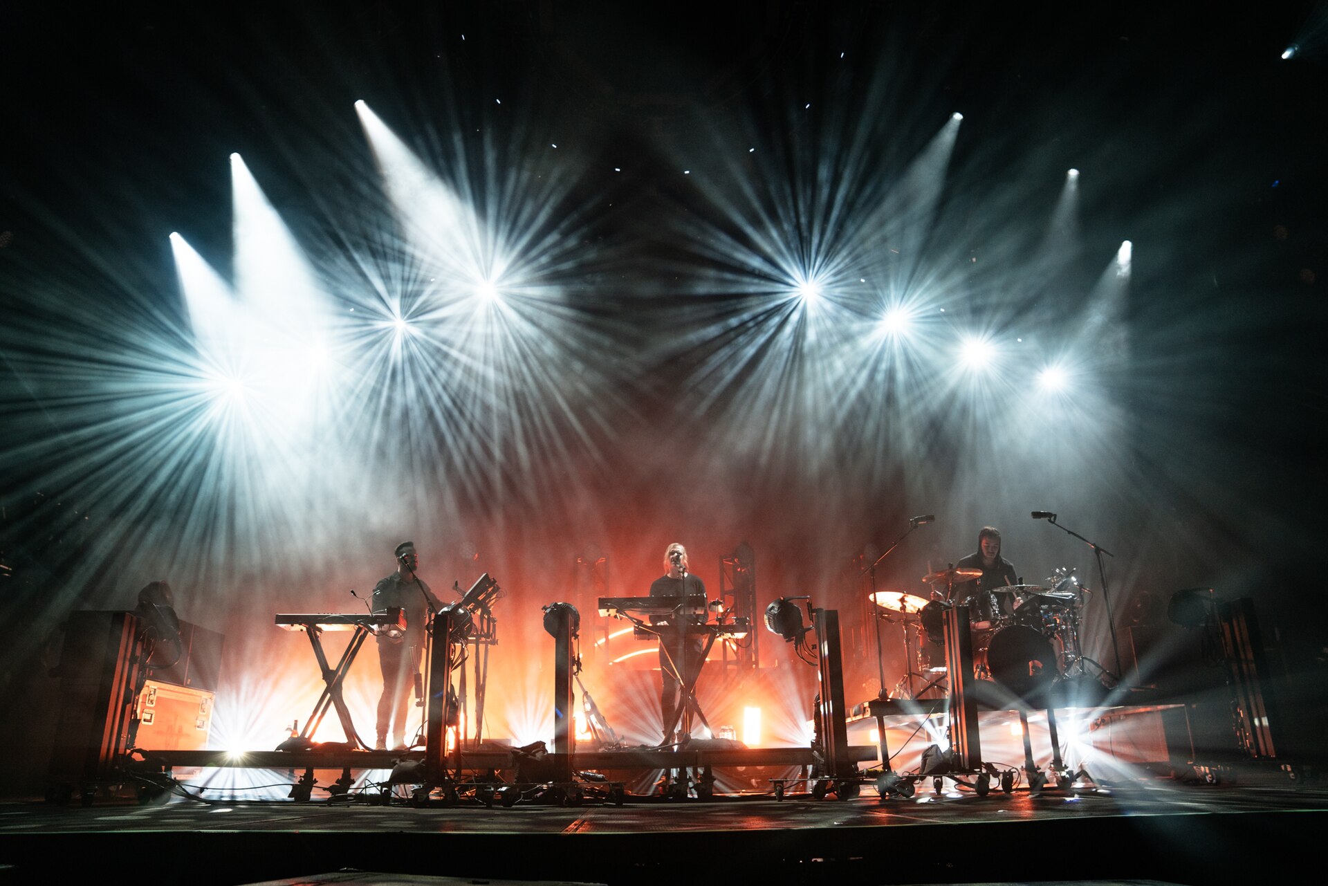 The three members of RUFUS DU SOL play a wide arena stage behind banks of keyboards and a drum kit, with starburst spotlights fanning out above them through orange haze.