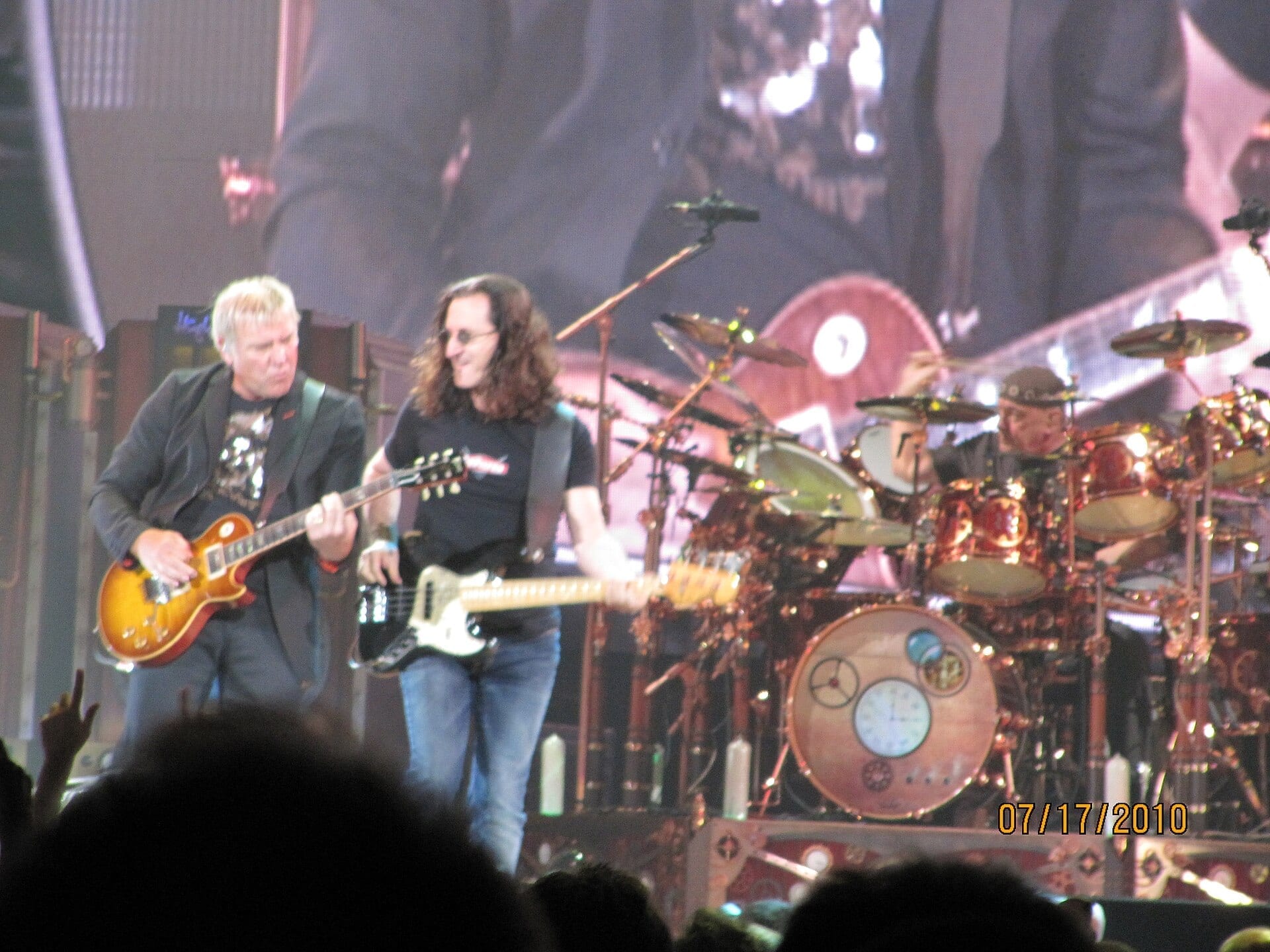 Rush guitarist Alex Lifeson and bassist-vocalist Geddy Lee perform side by side at the front of the stage, smiling, with drummer Neil Peart playing an elaborate steampunk-themed drum kit behind them.