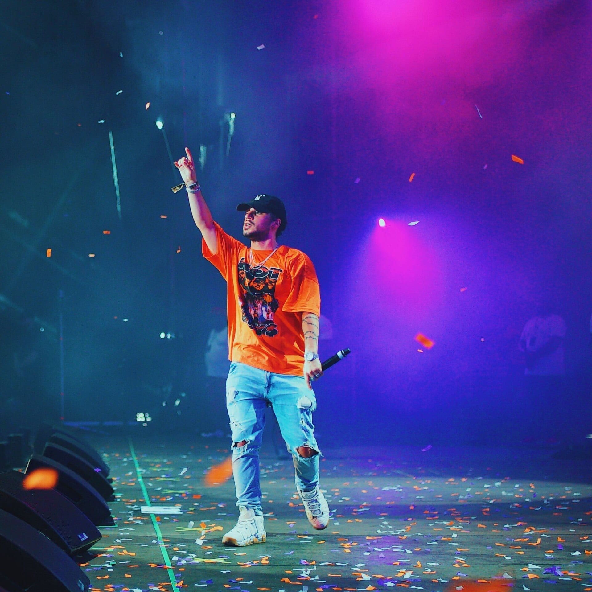 Russ performs on a smoky stage lit in pink and blue, one arm raised, wearing an orange graphic T-shirt, ripped jeans, and a baseball cap, as confetti falls around him.