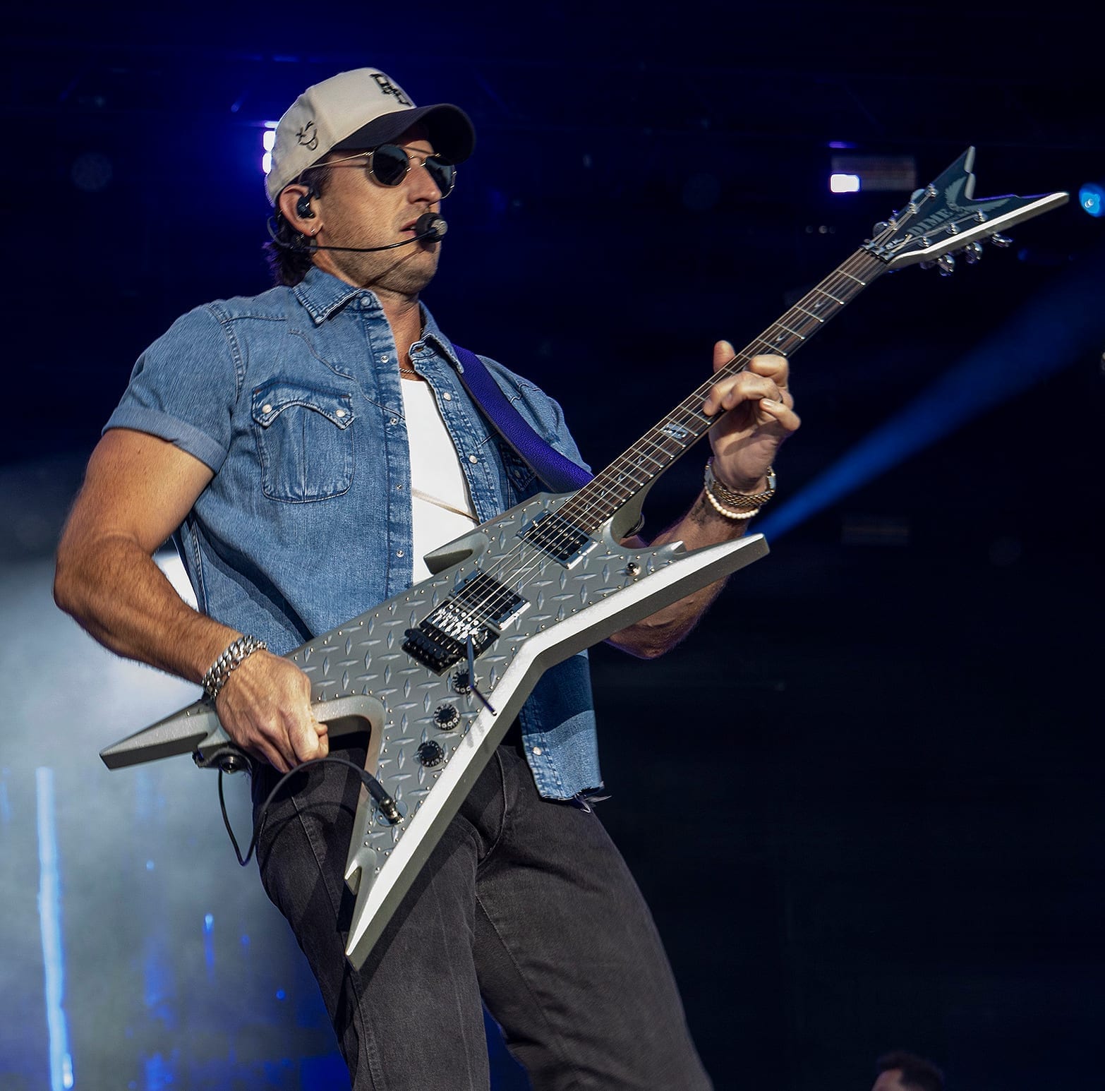 Russell Dickerson plays a silver flying-V electric guitar on stage, wearing a denim shirt, ball cap and sunglasses with a headset microphone.