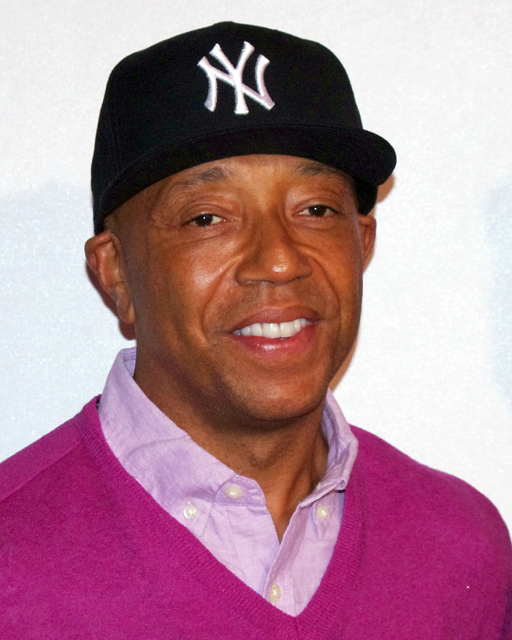 Russell Simmons smiles at the camera in a black New York Yankees cap and a magenta sweater over a lilac shirt.