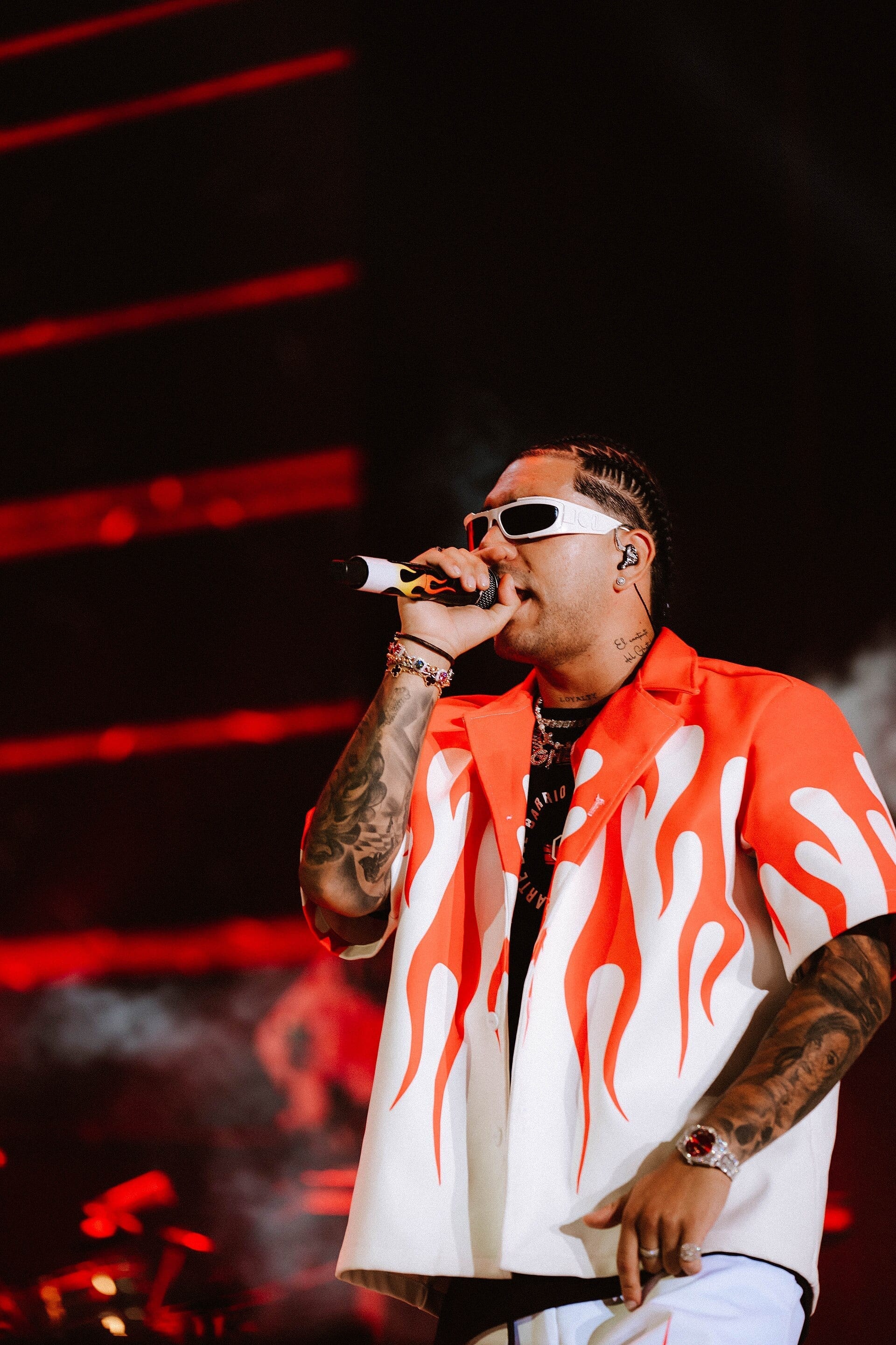 Ryan Castro sings into a flame-patterned microphone on stage, wearing a white and orange flame-print shirt and sunglasses, with tattoos visible on both arms.