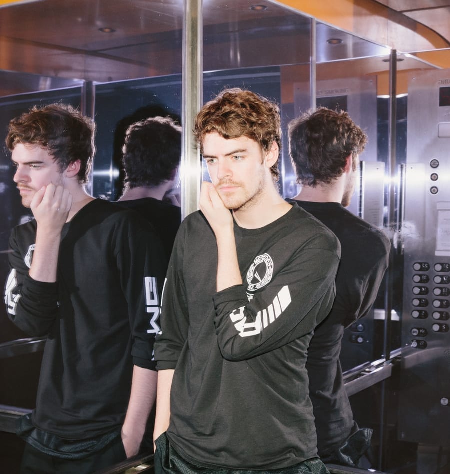 Ryan Hemsworth stands in a mirrored lift with his chin resting on one hand, in a black long-sleeved shirt, his reflection repeating on both sides.