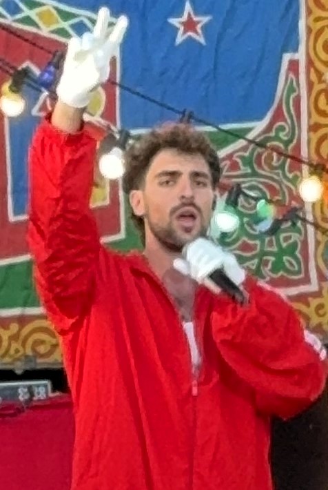 Saint Levant sings into a handheld microphone in a red tracksuit and white gloves, one hand raised, festoon lights behind him.