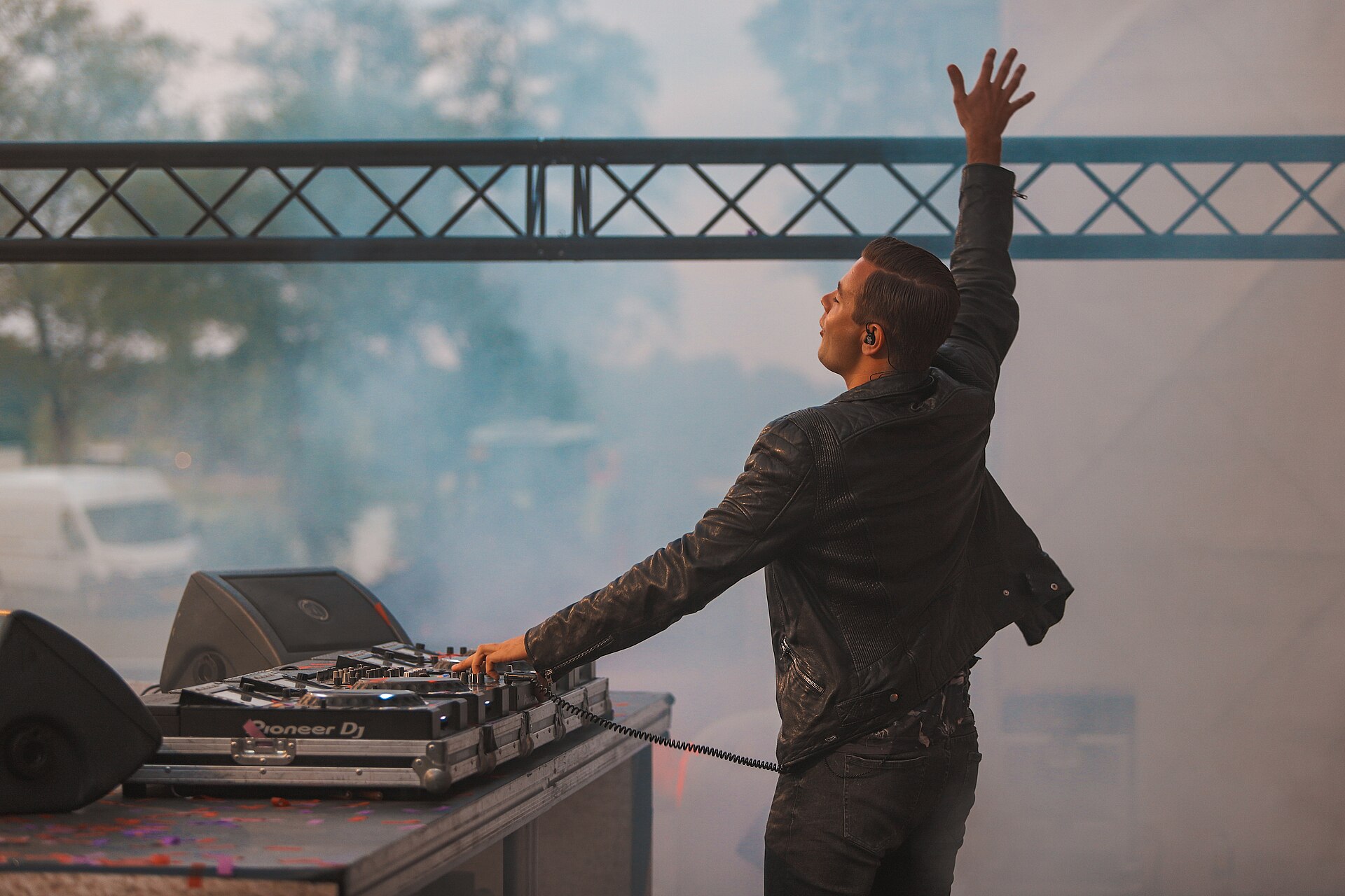 Sam Feldt stands behind a Pioneer DJ setup with one arm thrown in the air, smoke drifting across an outdoor stage.