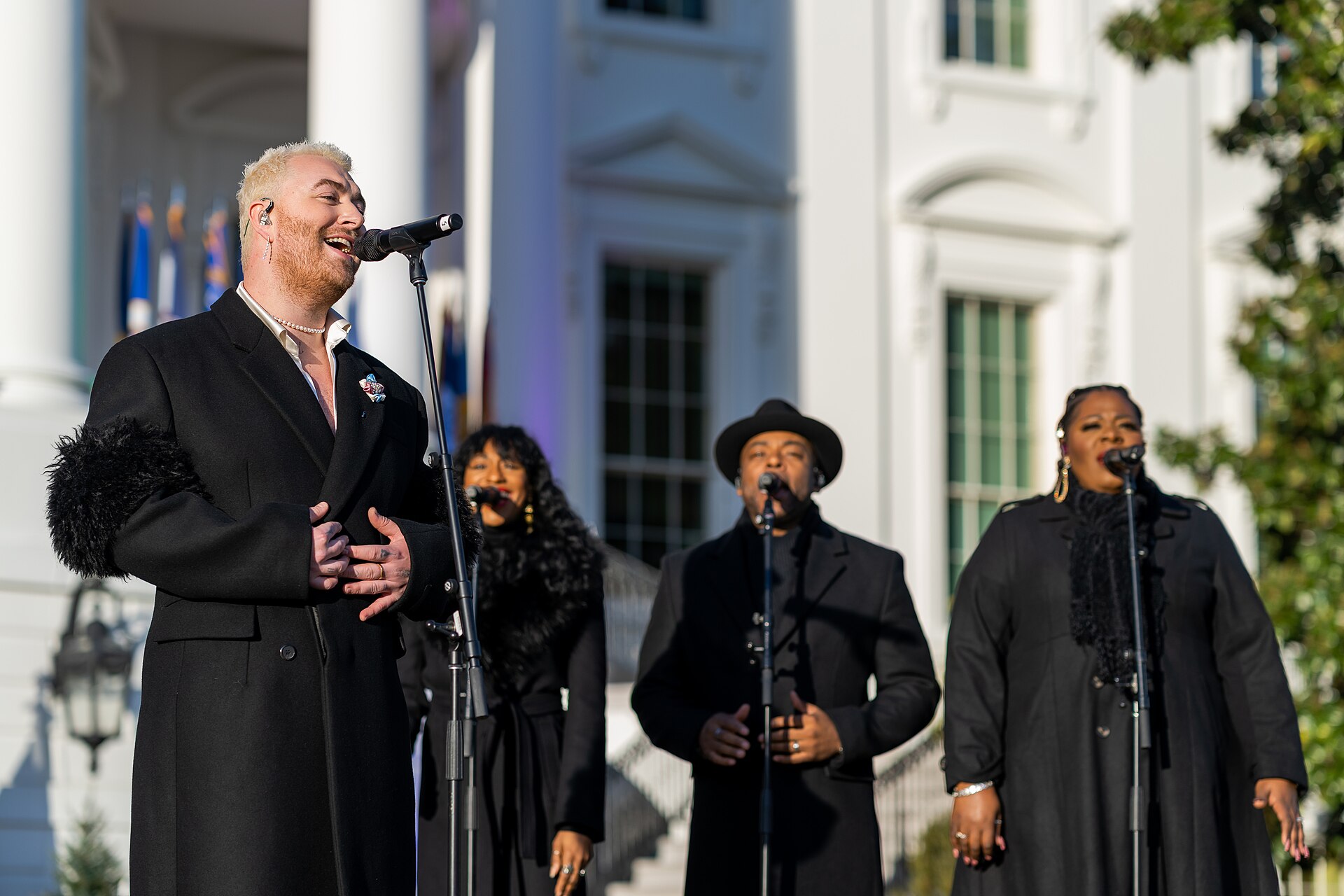 Sam Smith sings at a mic stand in a black coat with fur cuffs, three backing singers in black behind them and a white colonnade beyond.