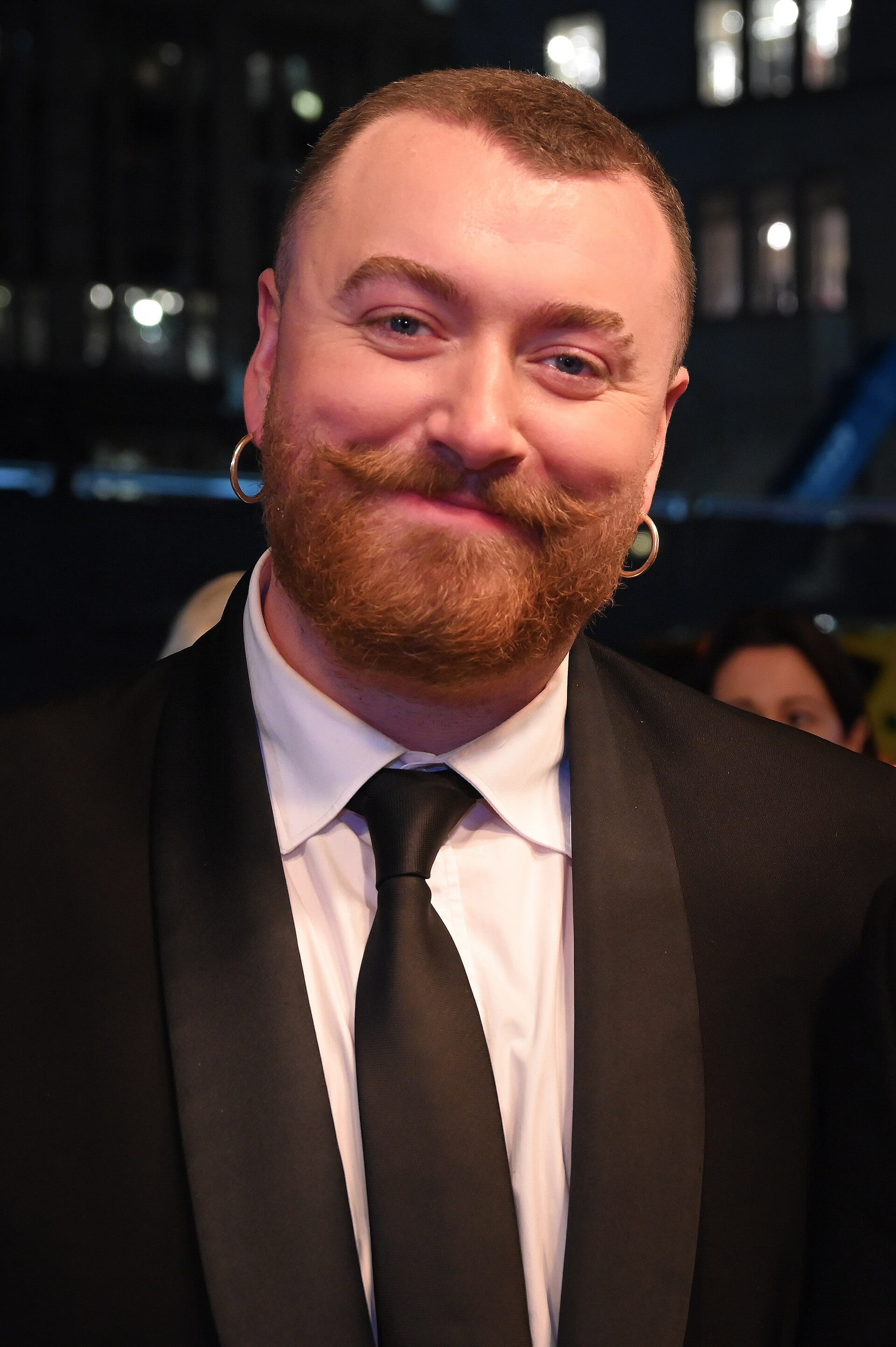 Sam Smith smiles at a red carpet event at night, wearing a black tuxedo with satin lapels, a white shirt and black tie, hoop earrings, and a ginger beard and mustache.