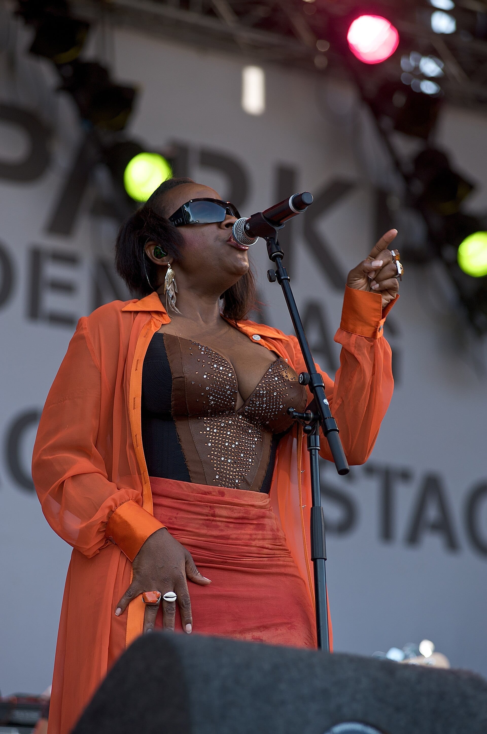 Sandra St. Victor sings into a stand microphone on an outdoor festival stage in dark sunglasses, a sheer orange duster over a beaded bodice, one hand raised with a finger pointed up.