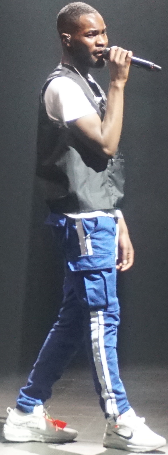 Dave raps into a microphone in profile, in a black gilet over a white T-shirt and blue tracksuit bottoms, against a plain dark backdrop.