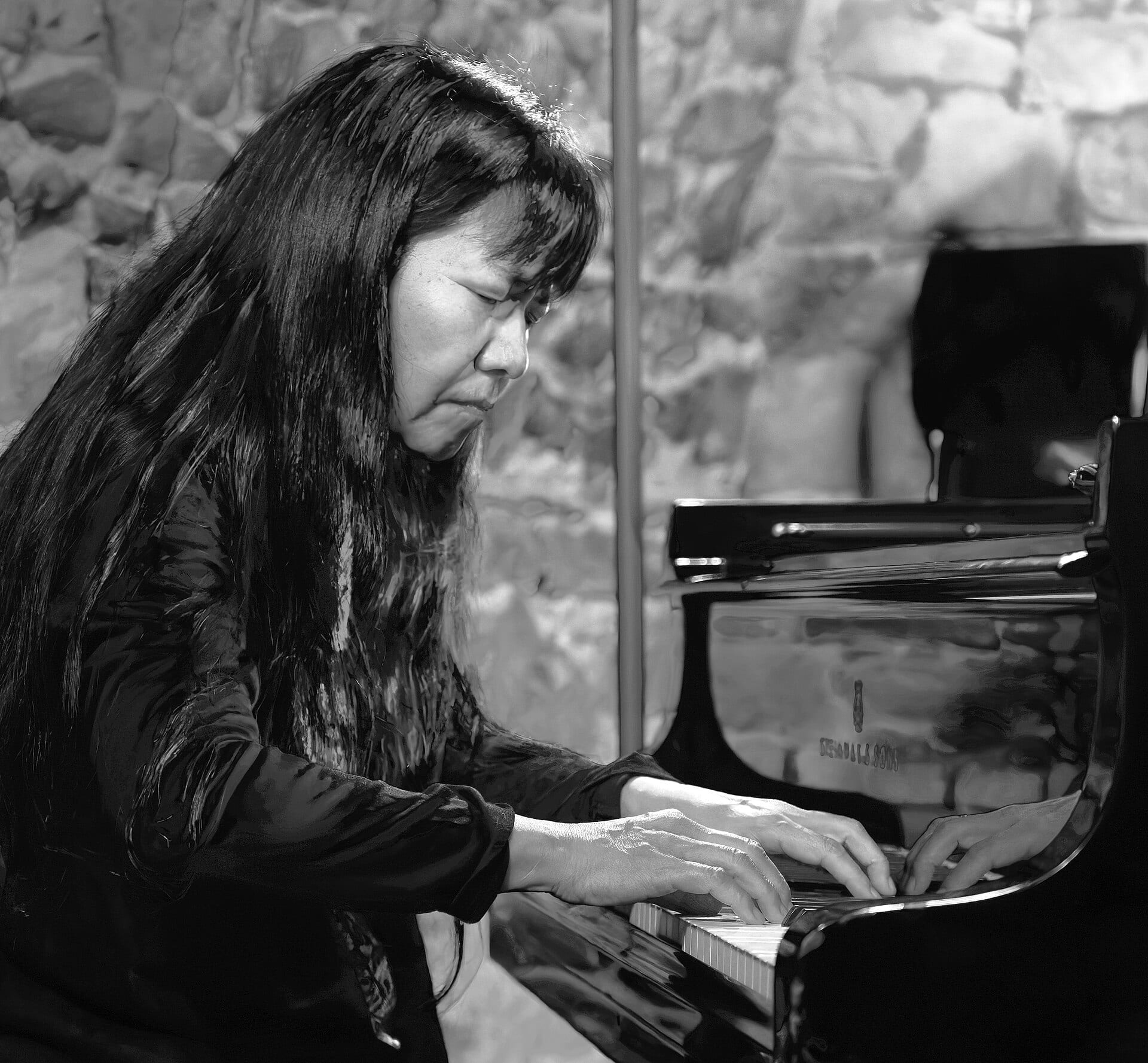 Satoko Fujii plays a grand piano with both hands on the keys, her long dark hair falling forward, in a black and white photograph.