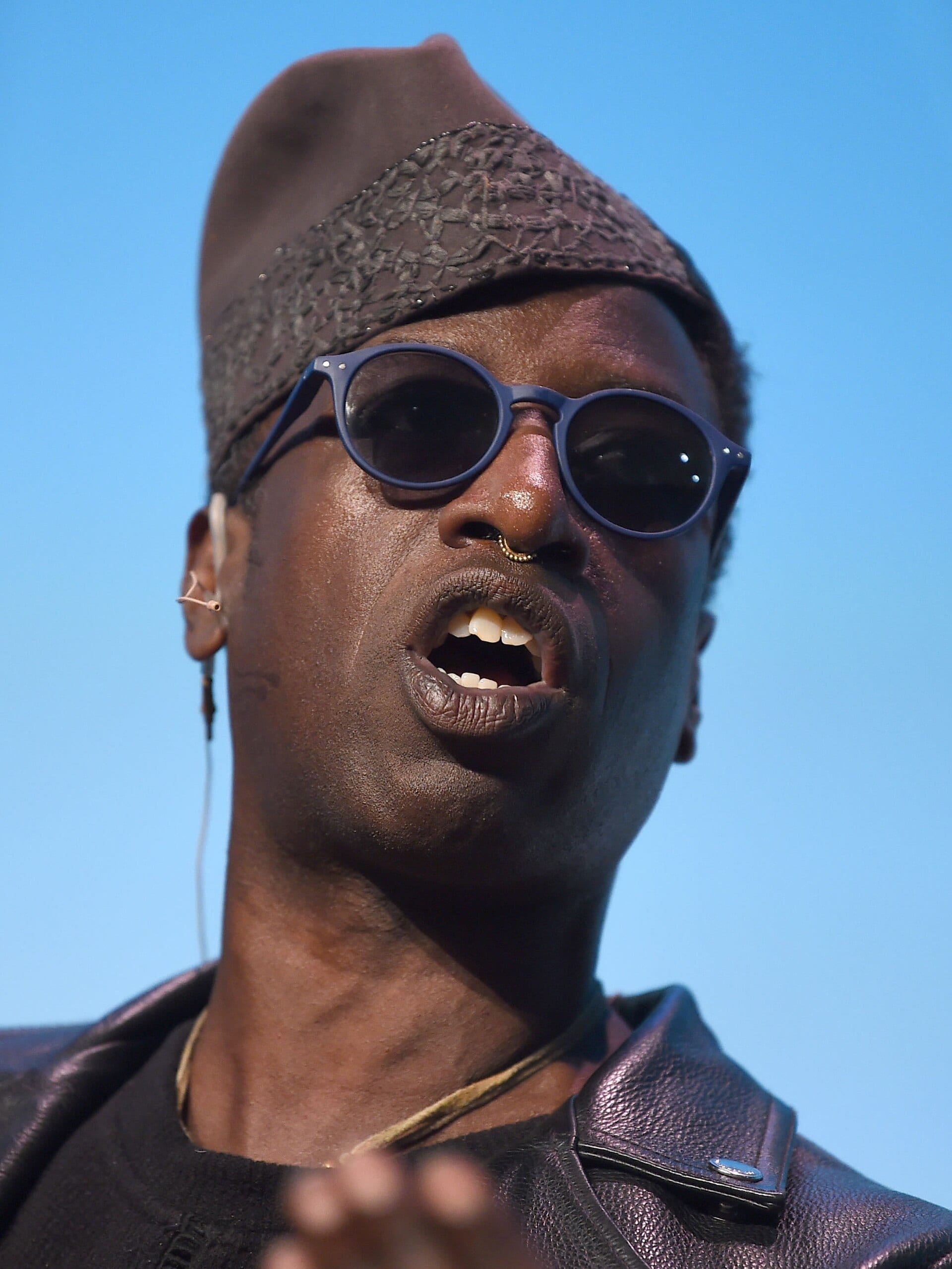 Saul Williams speaks mid-sentence against a clear blue sky, in round dark glasses, an embroidered cap and a black leather jacket.